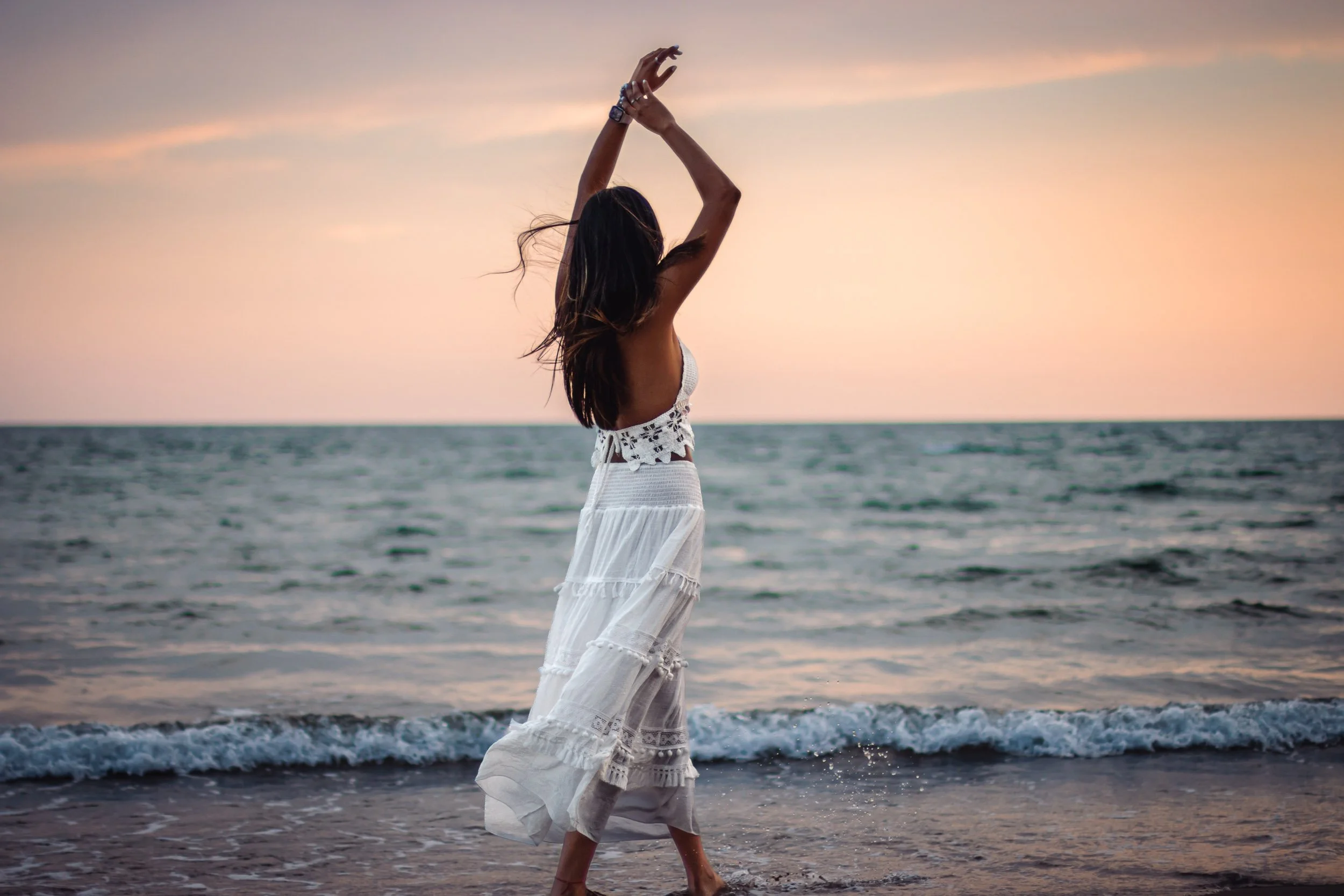 Back view of a woman in a white dress standing by the sea with her arms raised