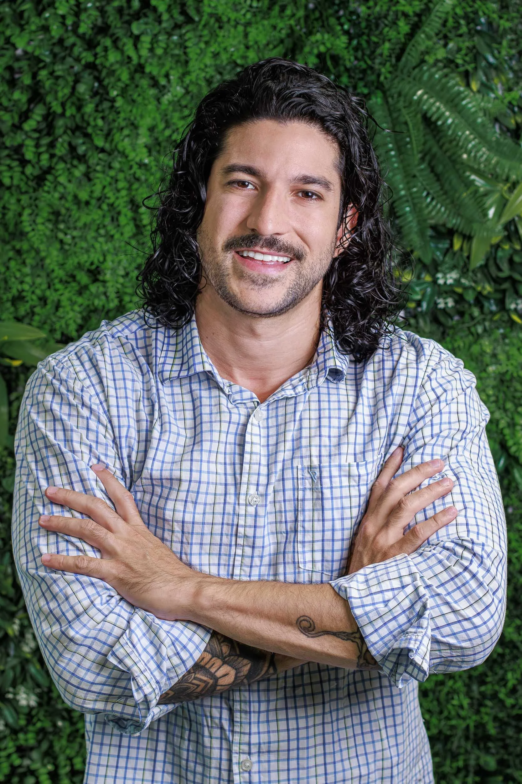 Professional headshot of a man with curly hair standing with arms crossed against a green plant wall