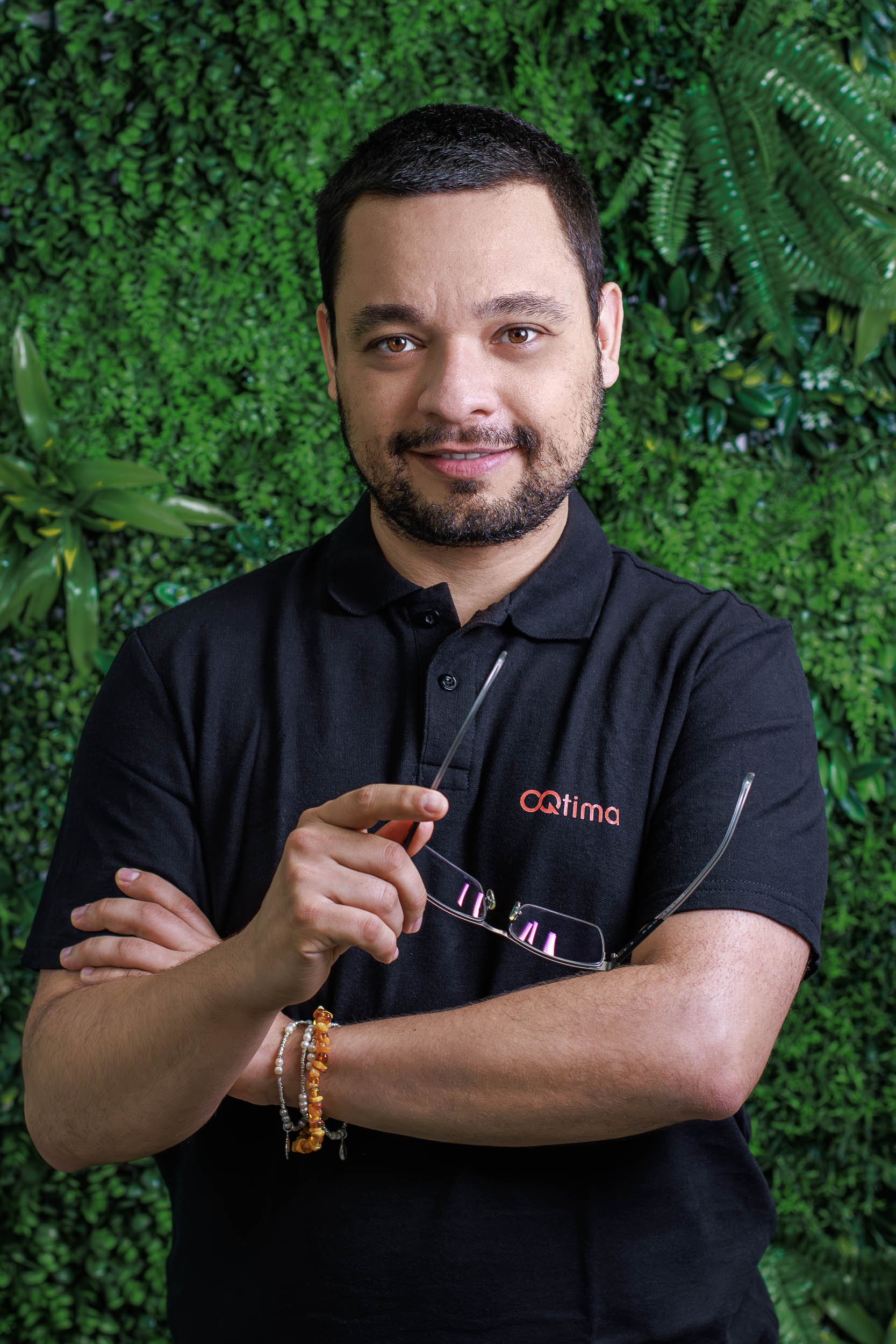 Professional headshot of a man holding glasses against a green plant wall