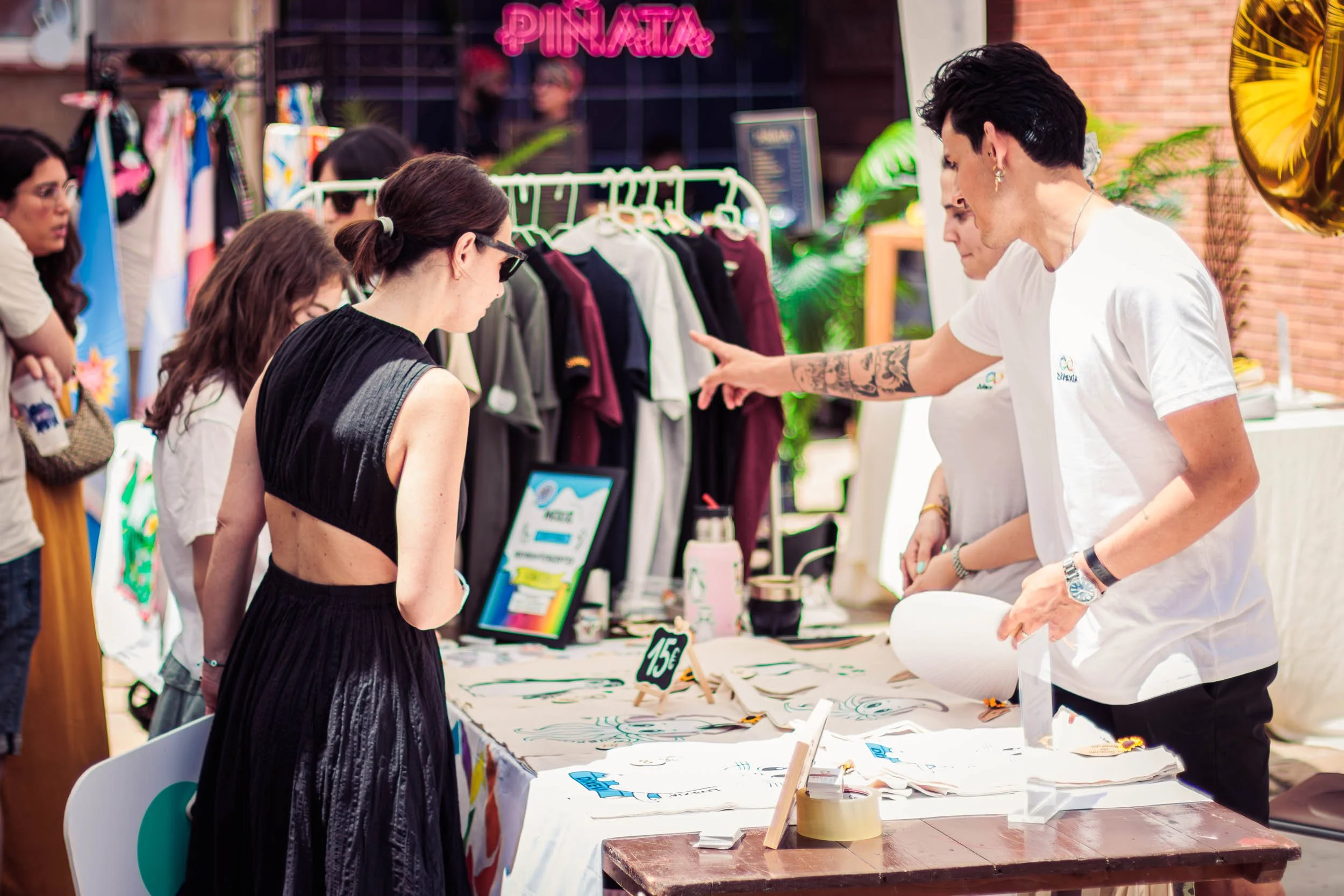 People gathered around a table at an indoor market event, looking at displayed items while a vendor in a white shirt gestures toward the stand.