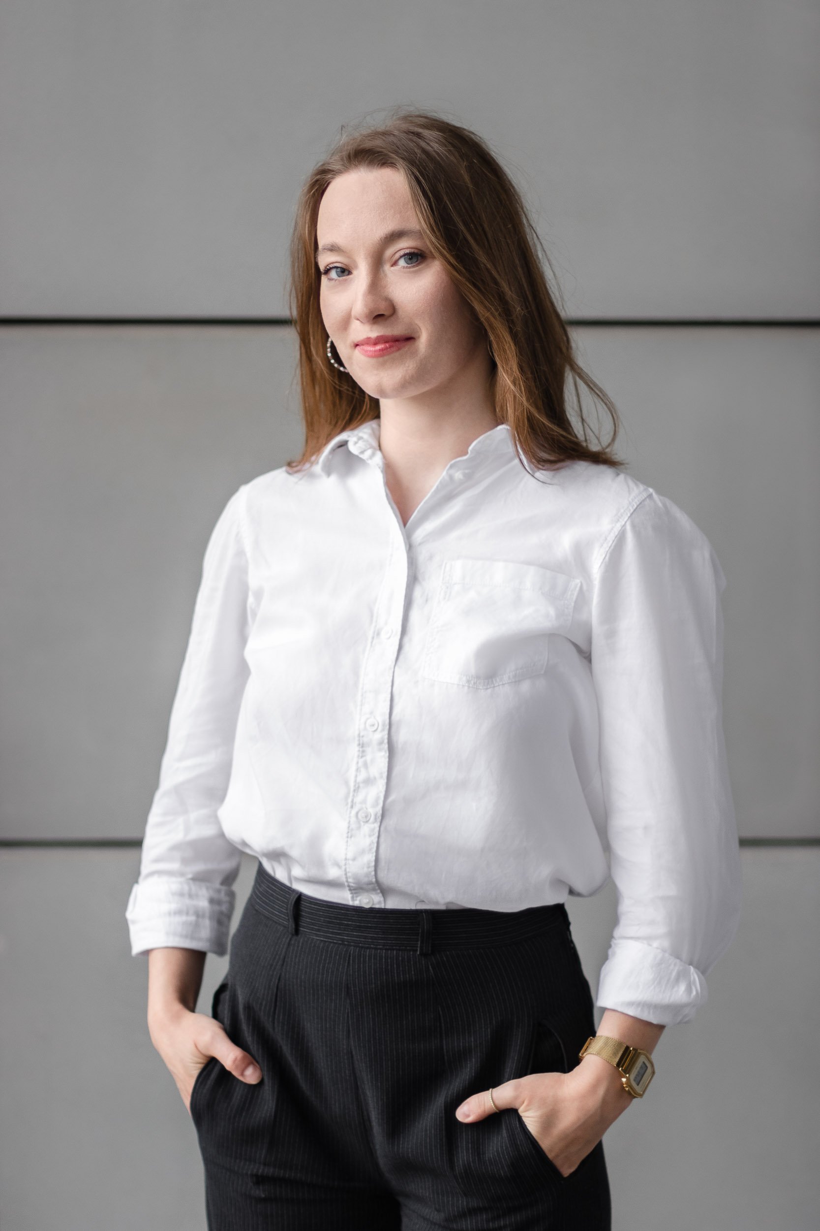 Full-length professional portrait of a woman standing with hands in her pockets against a grey wall