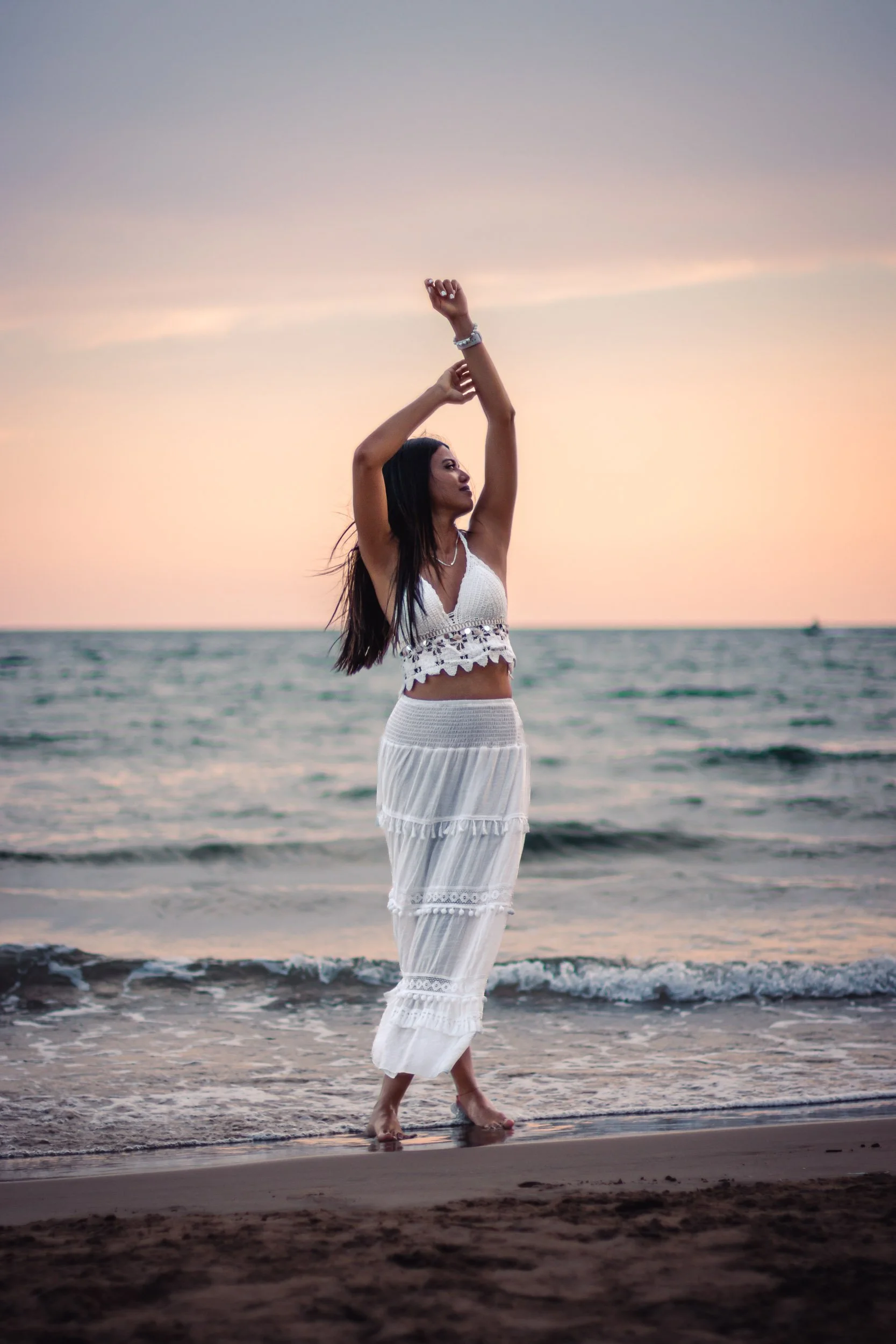 Full-length portrait of a woman standing on the beach with her arms raised at sunset