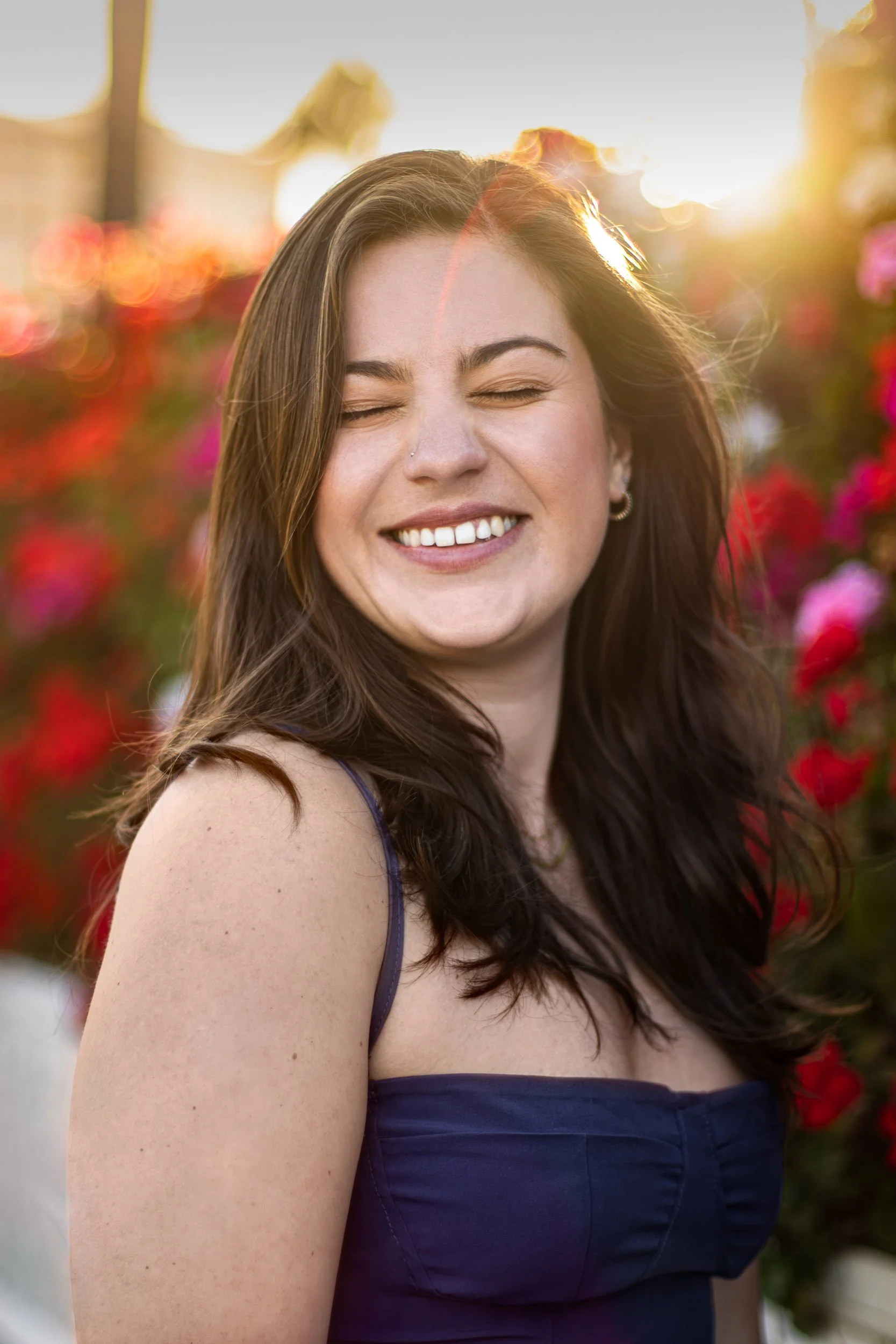 Woman in a dark blue strapless dress smiling with her eyes closed, standing outdoors in warm light with blurred flowers behind her.
