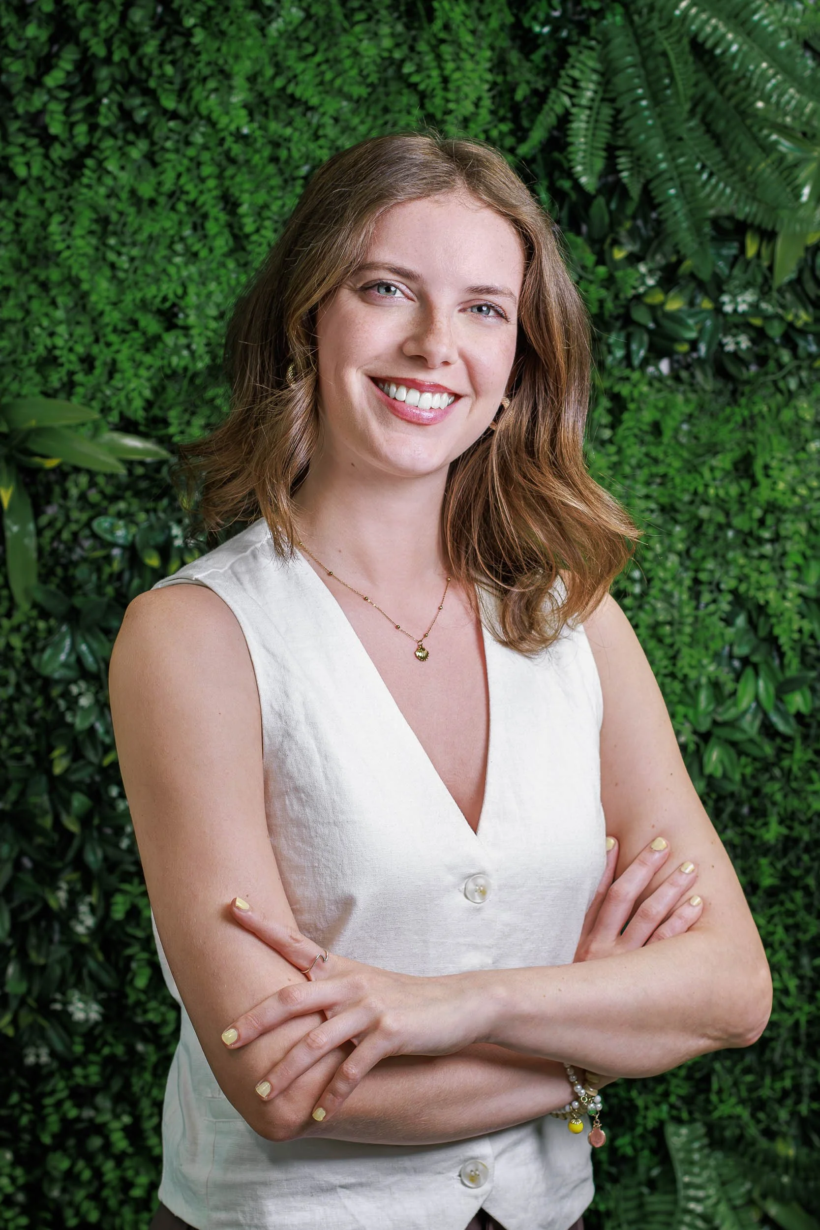Professional headshot of a smiling woman in a sleeveless top standing against a green plant wall