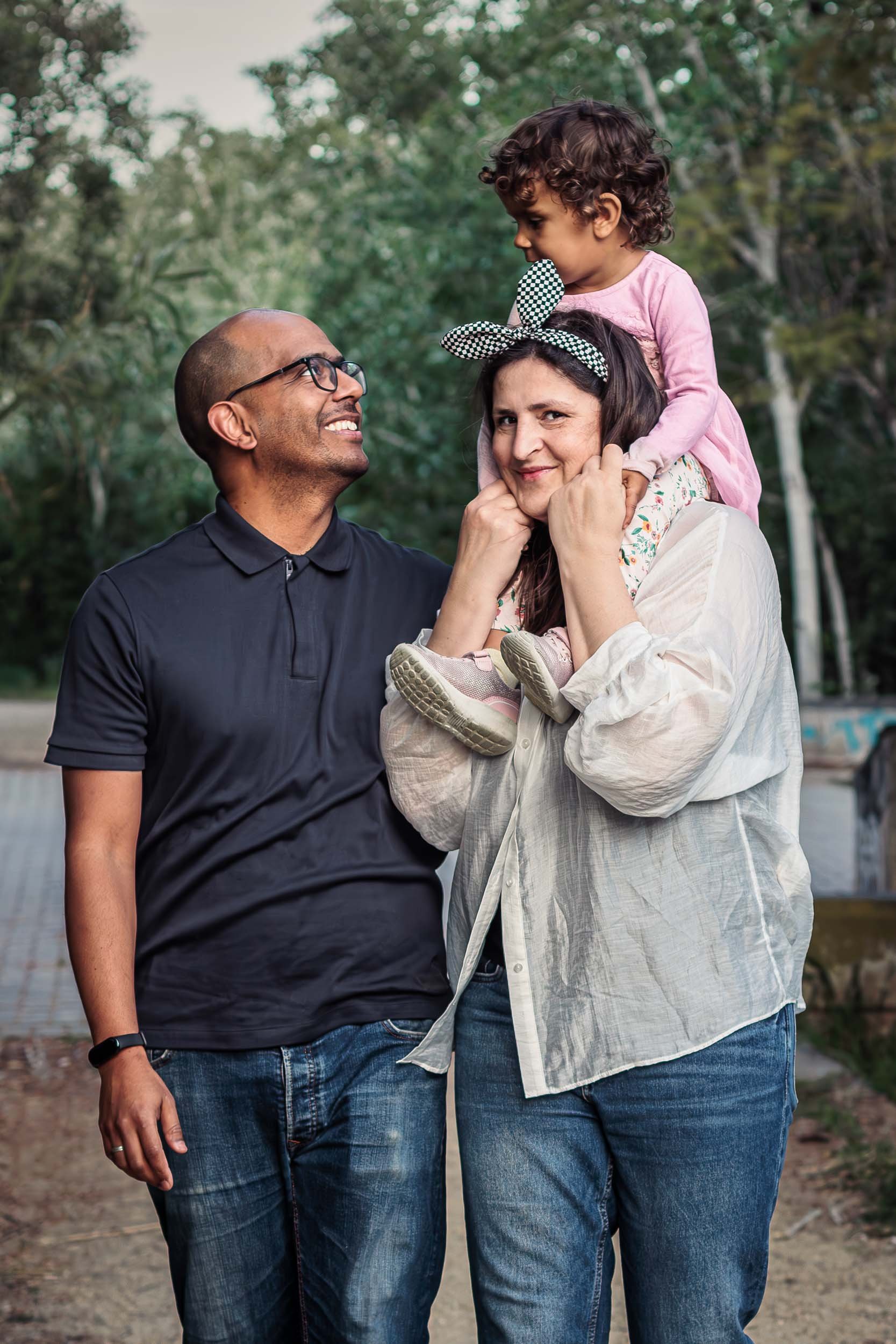 Portrait of two parents standing together with their daughter sitting on her mother's shoulders