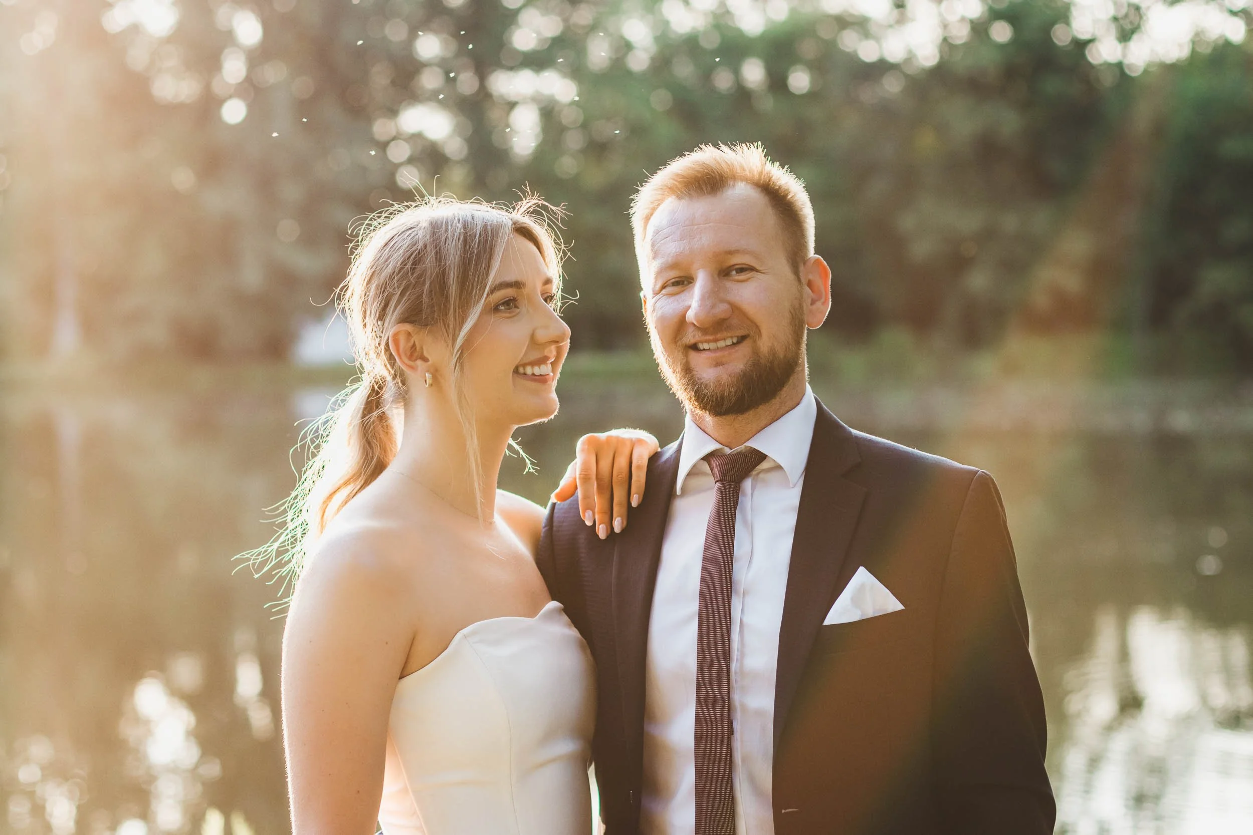 Wedding portrait of bride and groom smiling at each other while standing by the lake