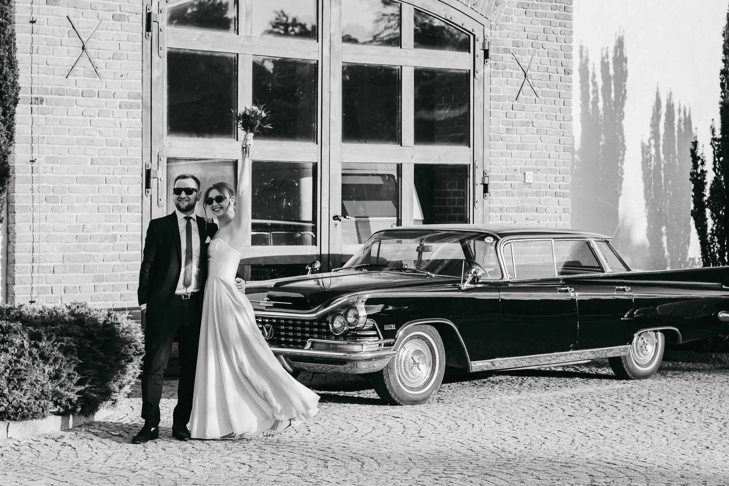 Black and white wedding portrait of the bride and groom standing beside a vintage black car