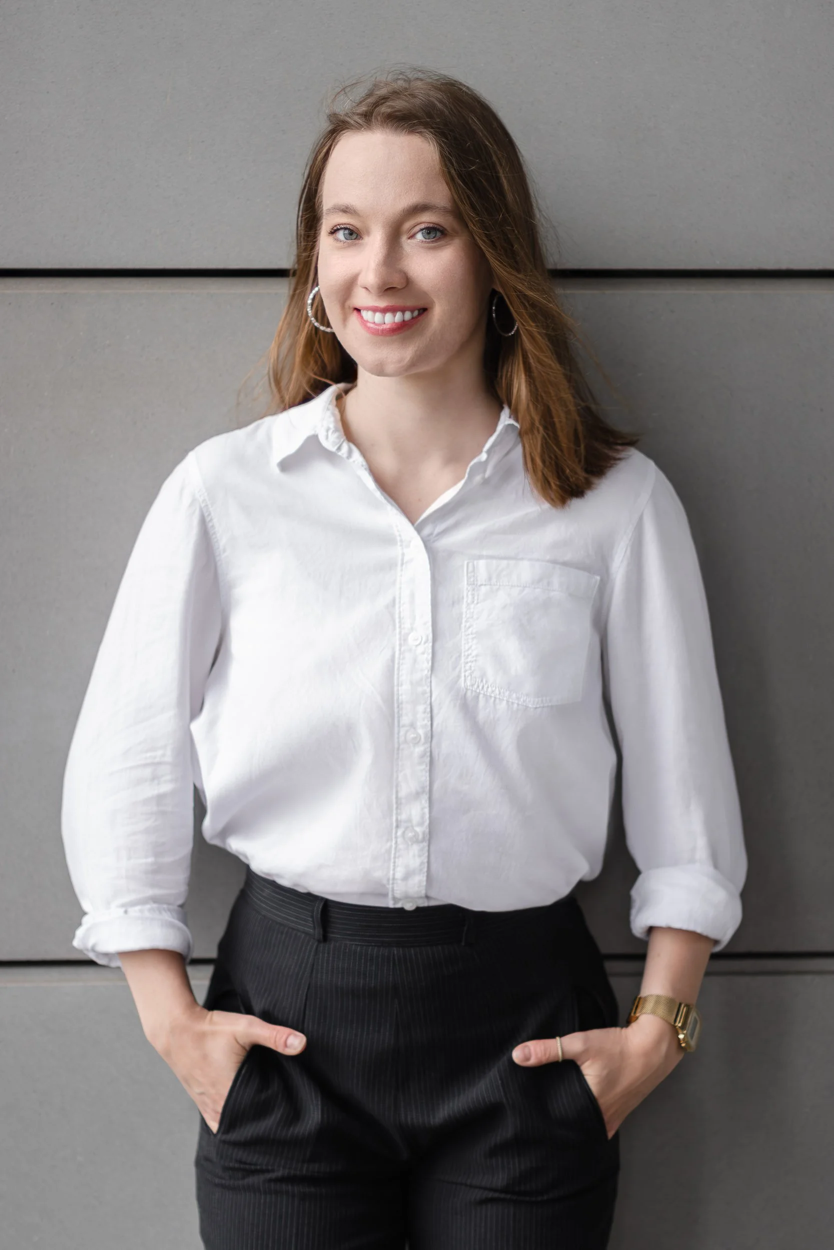 Full-length professional portrait of a woman standing with hands in her pockets against a grey wall