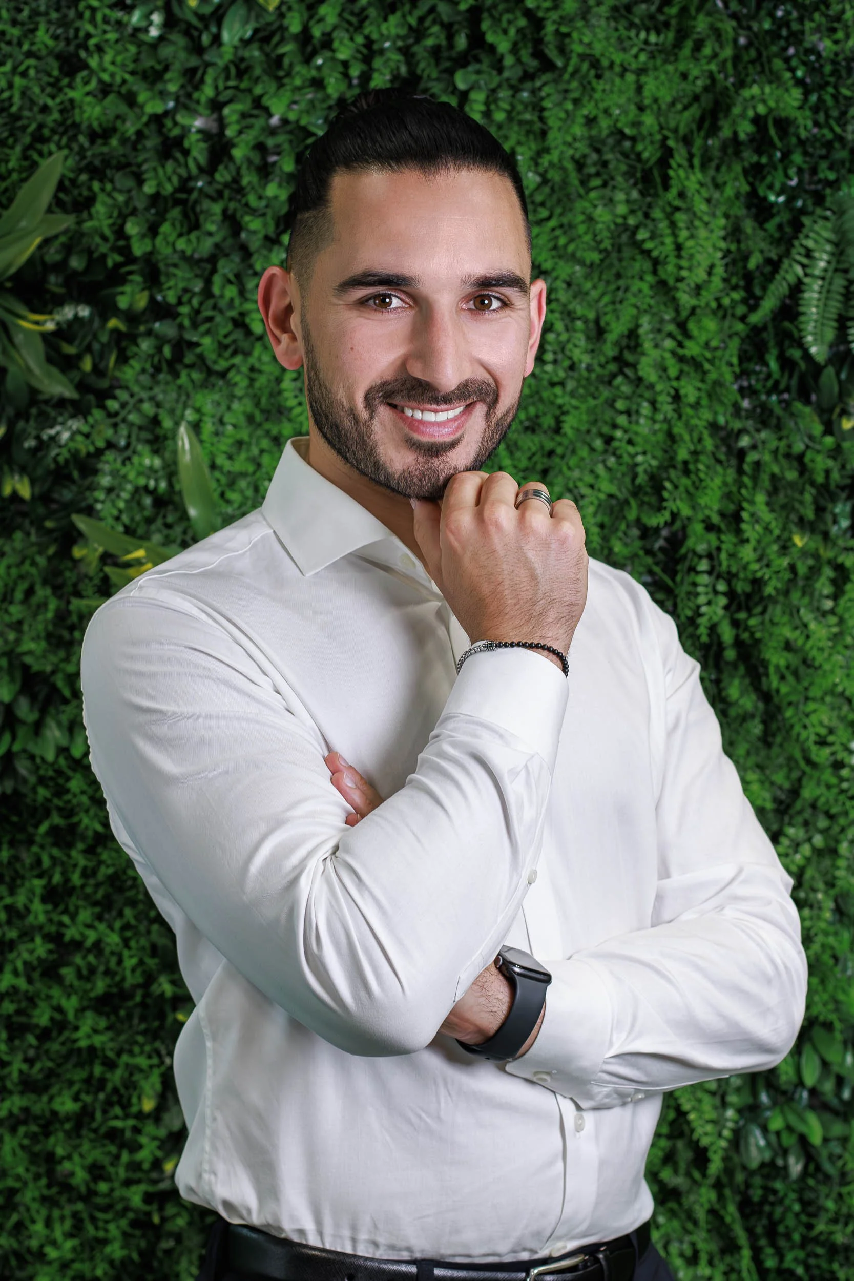 Professional headshot of a man in a white shirt standing with arms crossed against a green plant wall