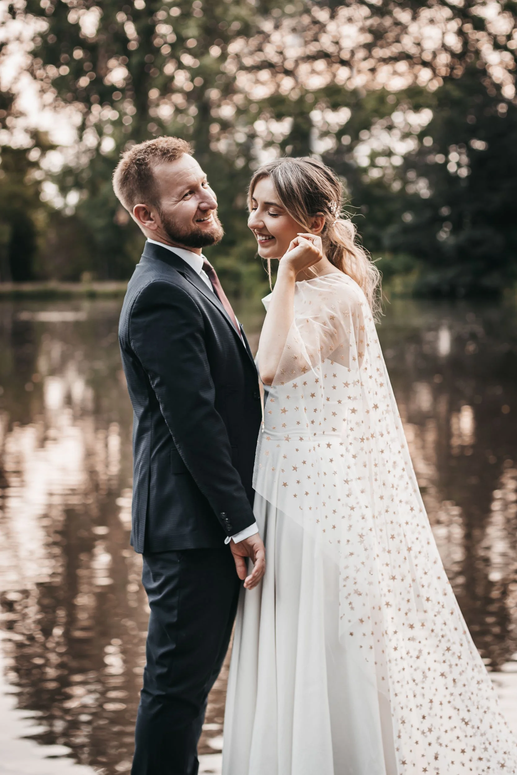 Full-length wedding portrait of the bride and groom standing by the lake with the bride's veil visible