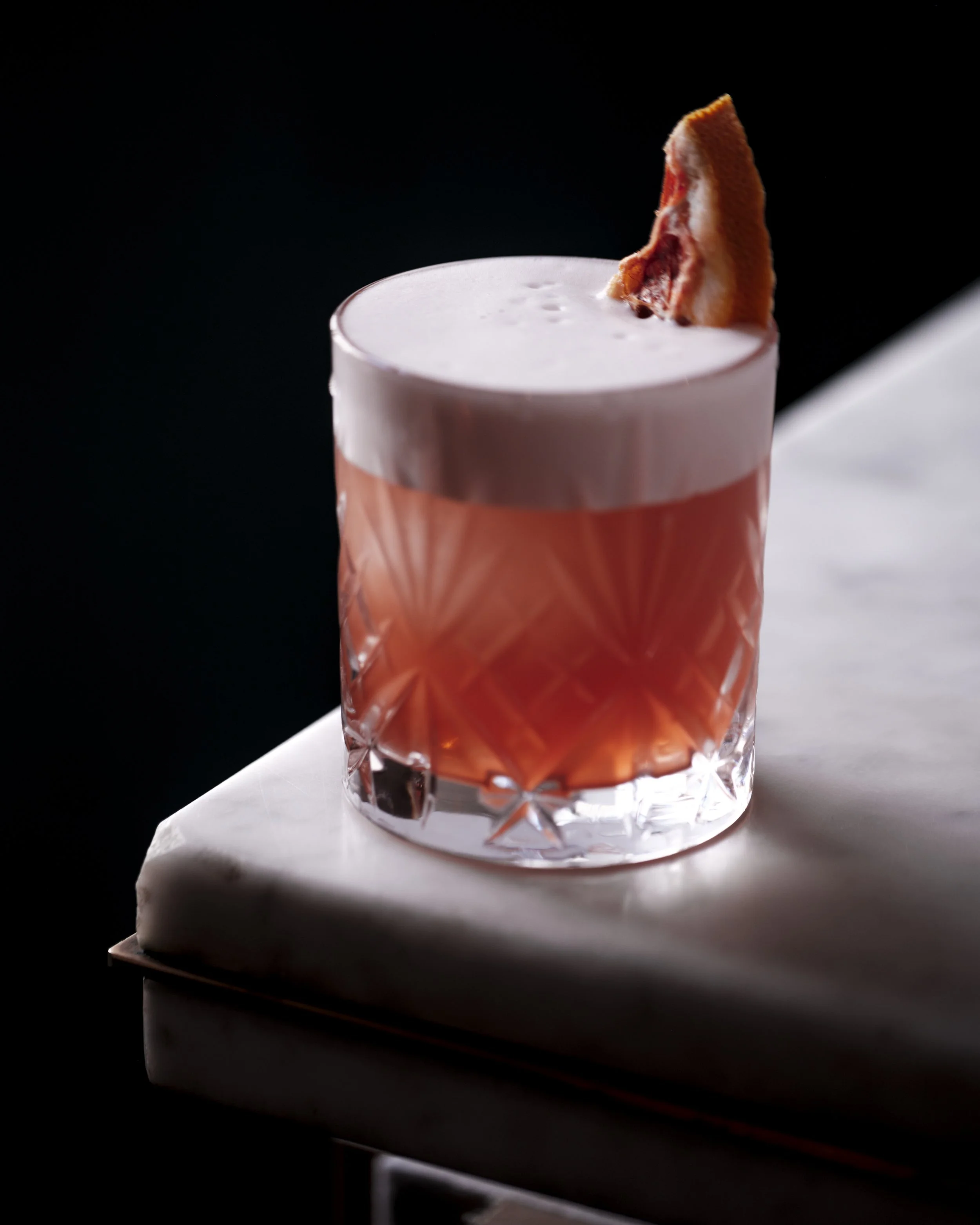 A cocktail with a pink frothy top and a slice of bacon garnish in a faceted glass on a white marble surface against a black background.