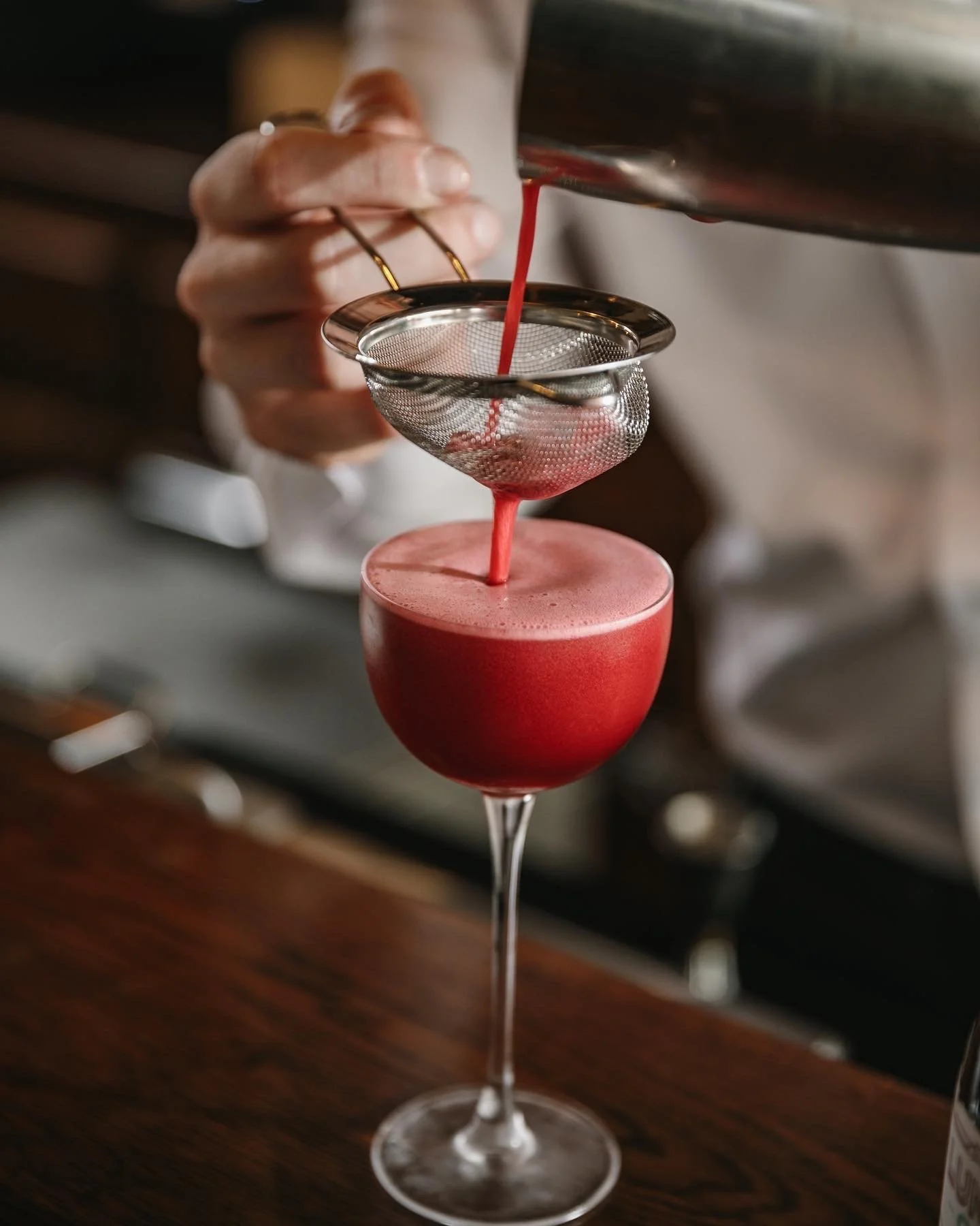 A bartender pours red cocktail through a fine mesh strainer into a pink frothy cocktail in a coupe glass, with a layered presentation involving a metal shaker and strainer.