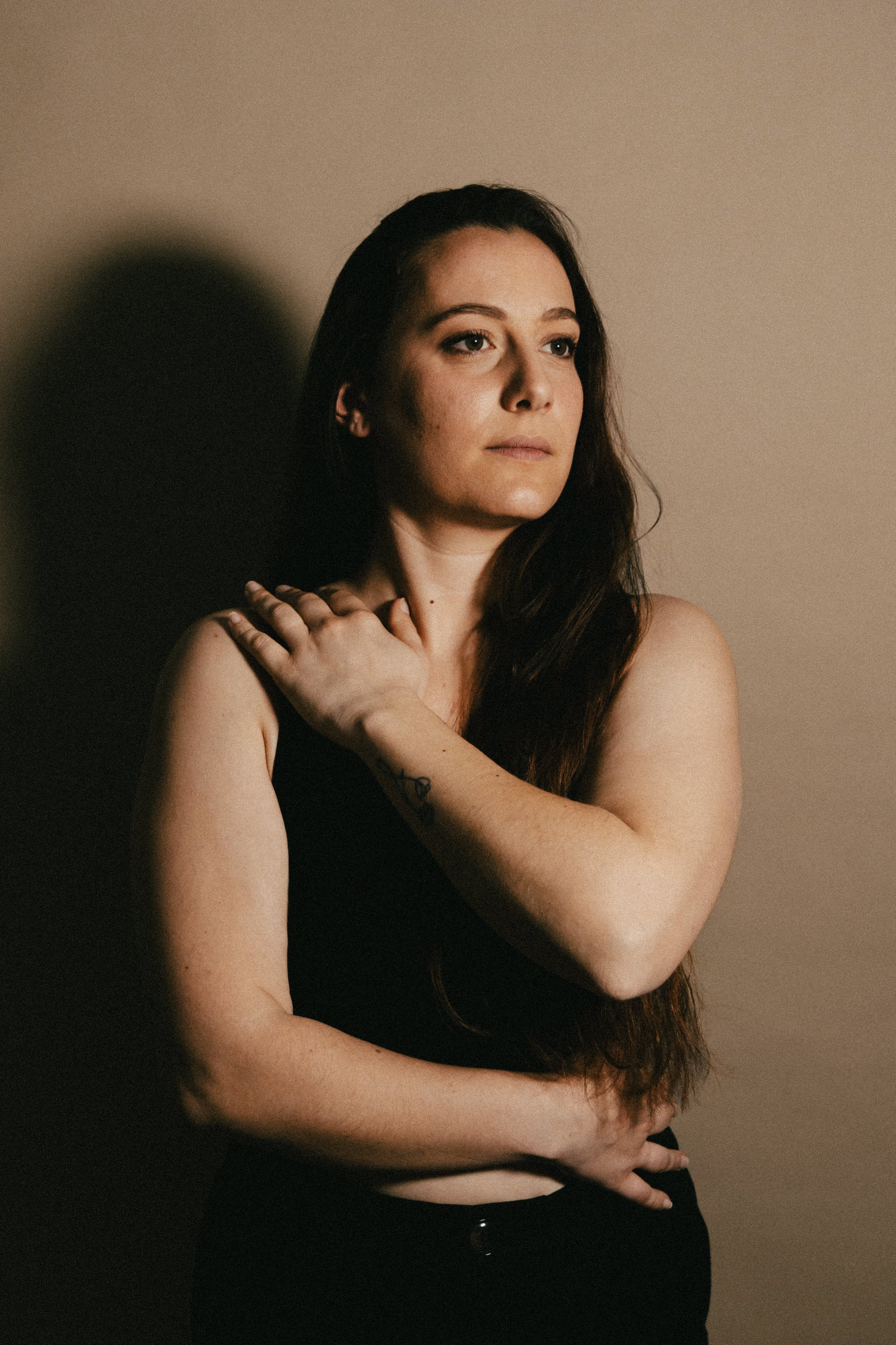 Une femme aux cheveux longs, portant un haut noir, pose contre un mur beige avec une ombre sombre derrière elle. Elle regarde au loin, la main gauche sur son épaule et la main droite sur son ventre.