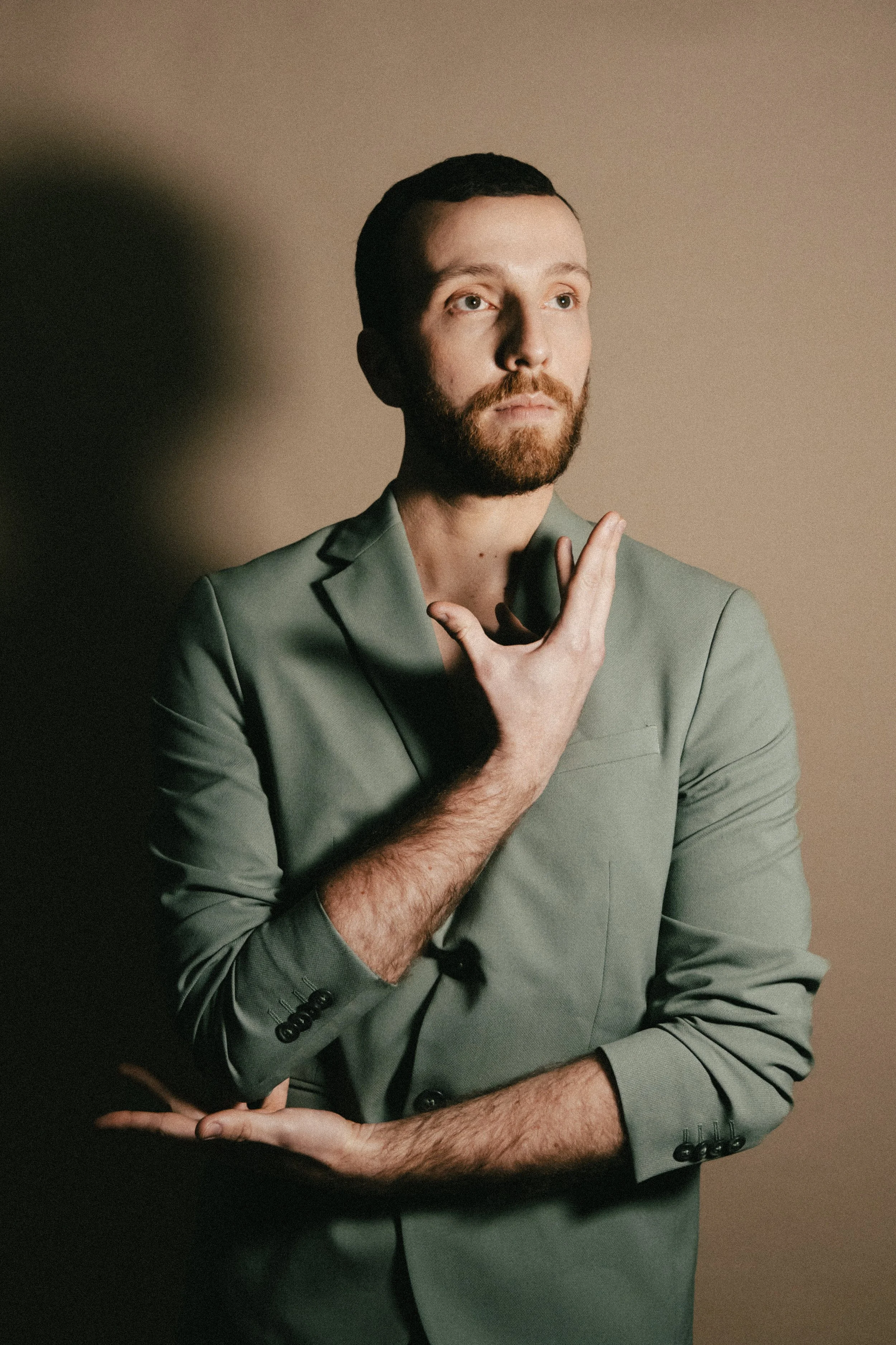 Un homme avec une barbe et des cheveux courts, portant un costume gris, pose avec une main près du visage contre un fond beige, avec un éclairage dramatique.