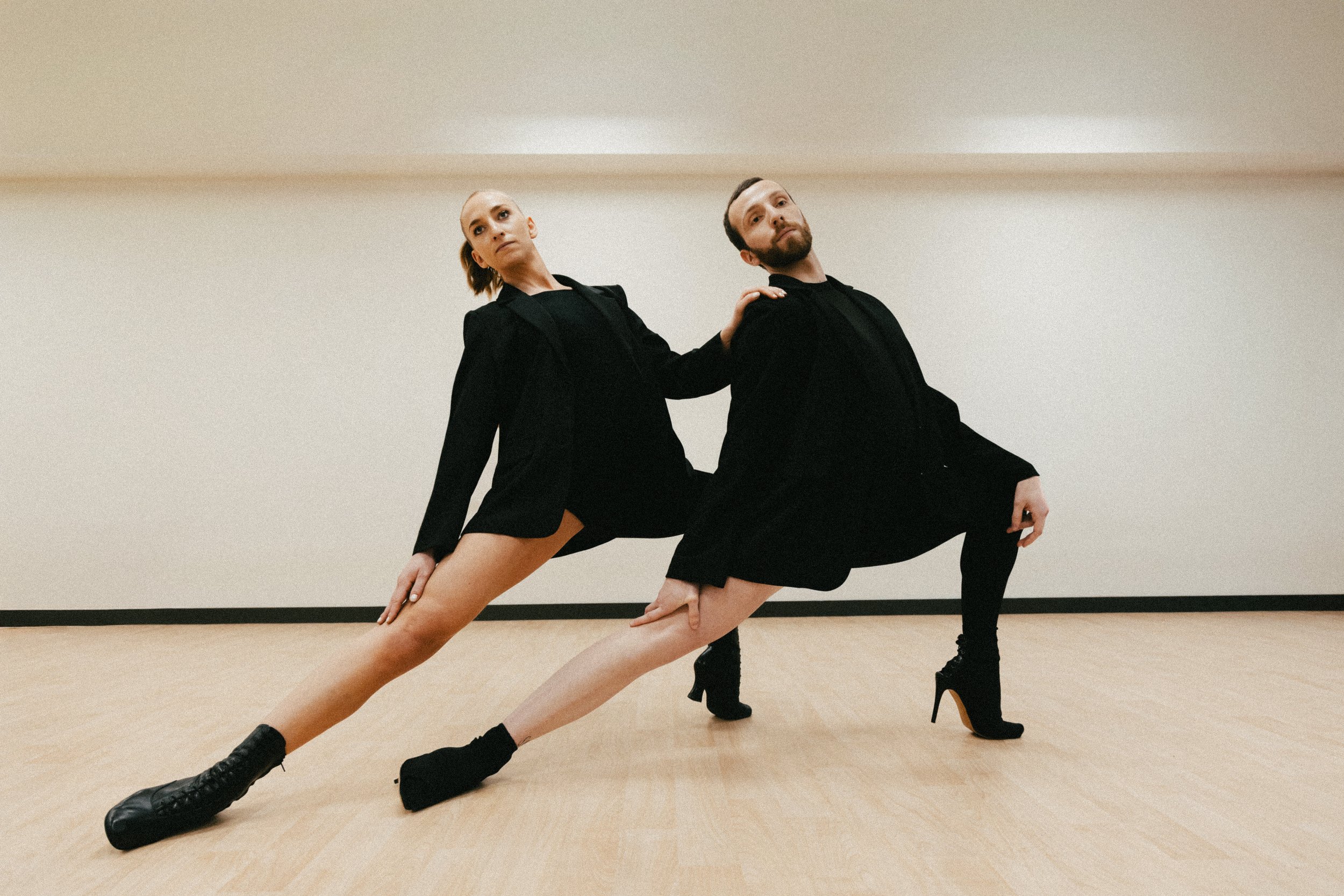 Deux danseurs en tenue noire en position de danse moderne ou contemporaine dans une pièce vide avec un sol en bois clair et un mur blanc. Ils sont accroupis, avec une jambe étendue en arrière, et une main posée sur leur genou ou leur cuisse pour équilibrer.