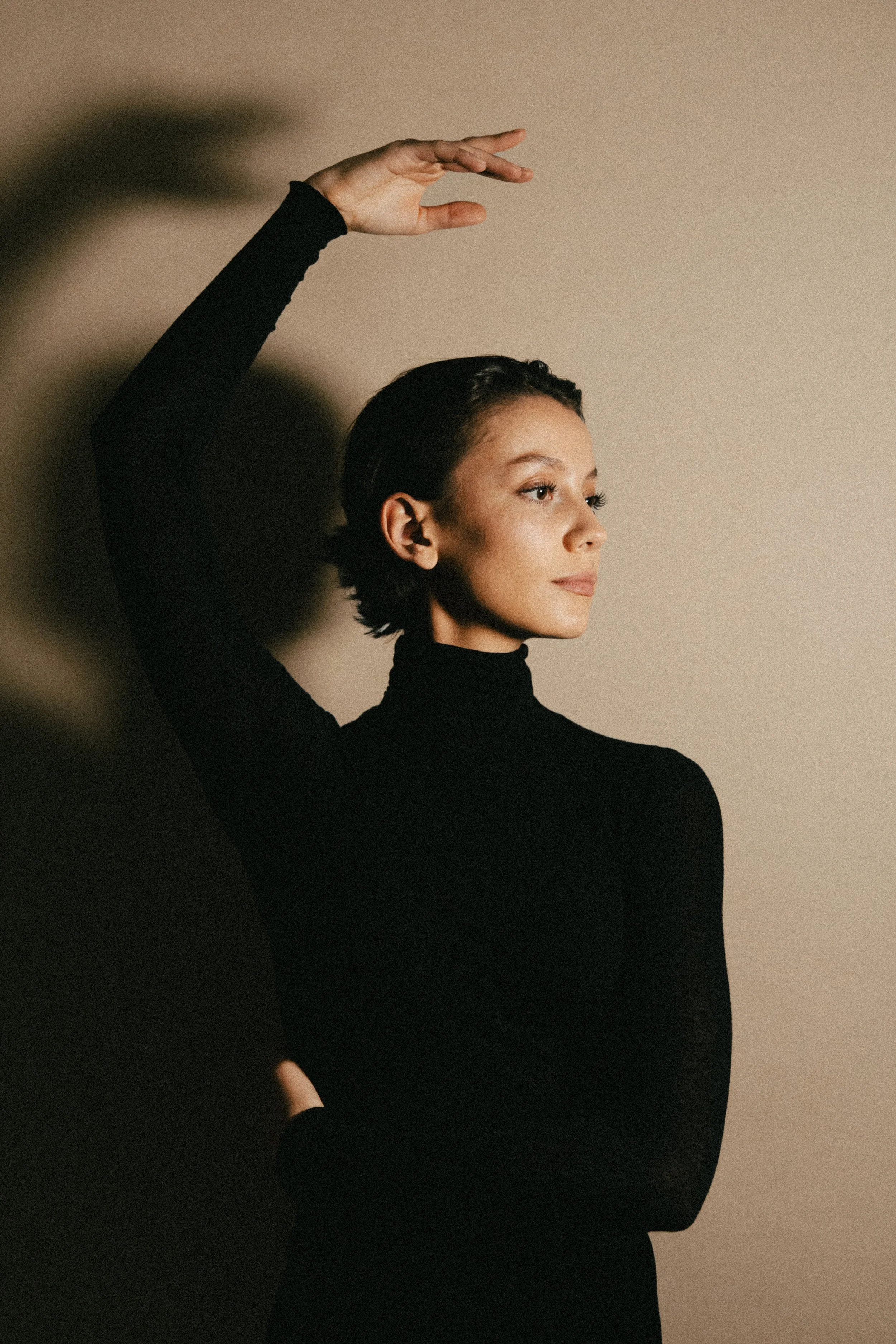 Femme avec les cheveux courts portant un haut à col roulé noir, posant contre un mur beige clair, levant une main au-dessus de sa tête, regardant vers la droite.