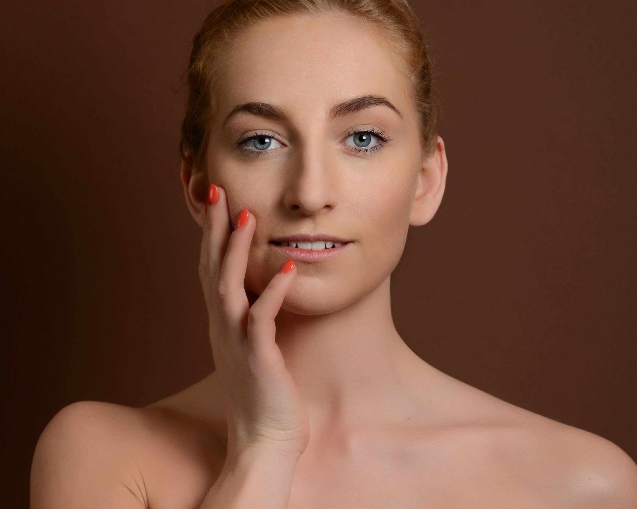 Portrait d'une femme avec peau claire, cheveux roux, regard bleu, touchant son visage avec ses mains, fond marron.