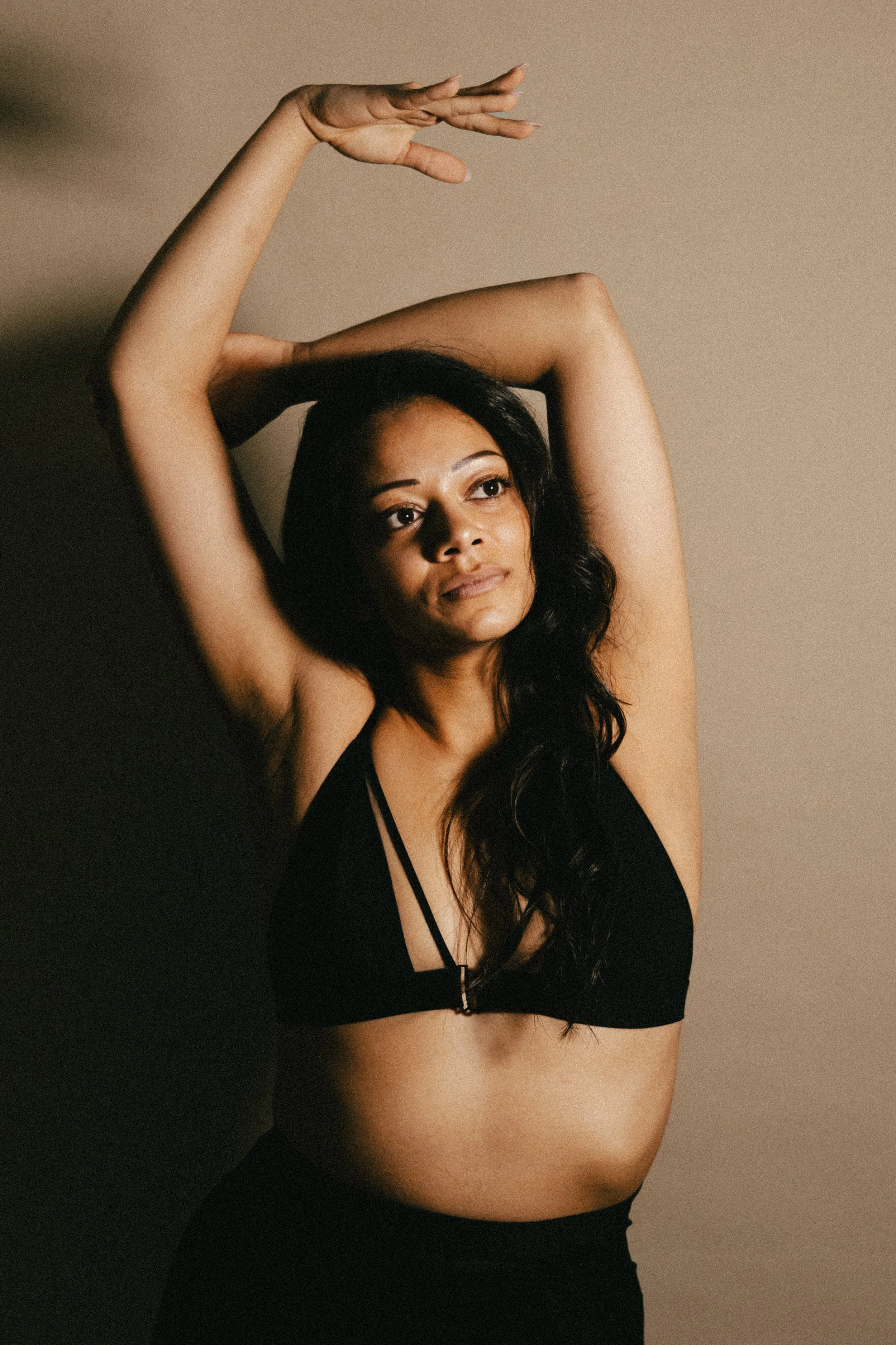 Une femme aux cheveux noirs longs pose avec ses bras levés au-dessus de sa tête, portant un haut noir sans manches, contre un fond beige