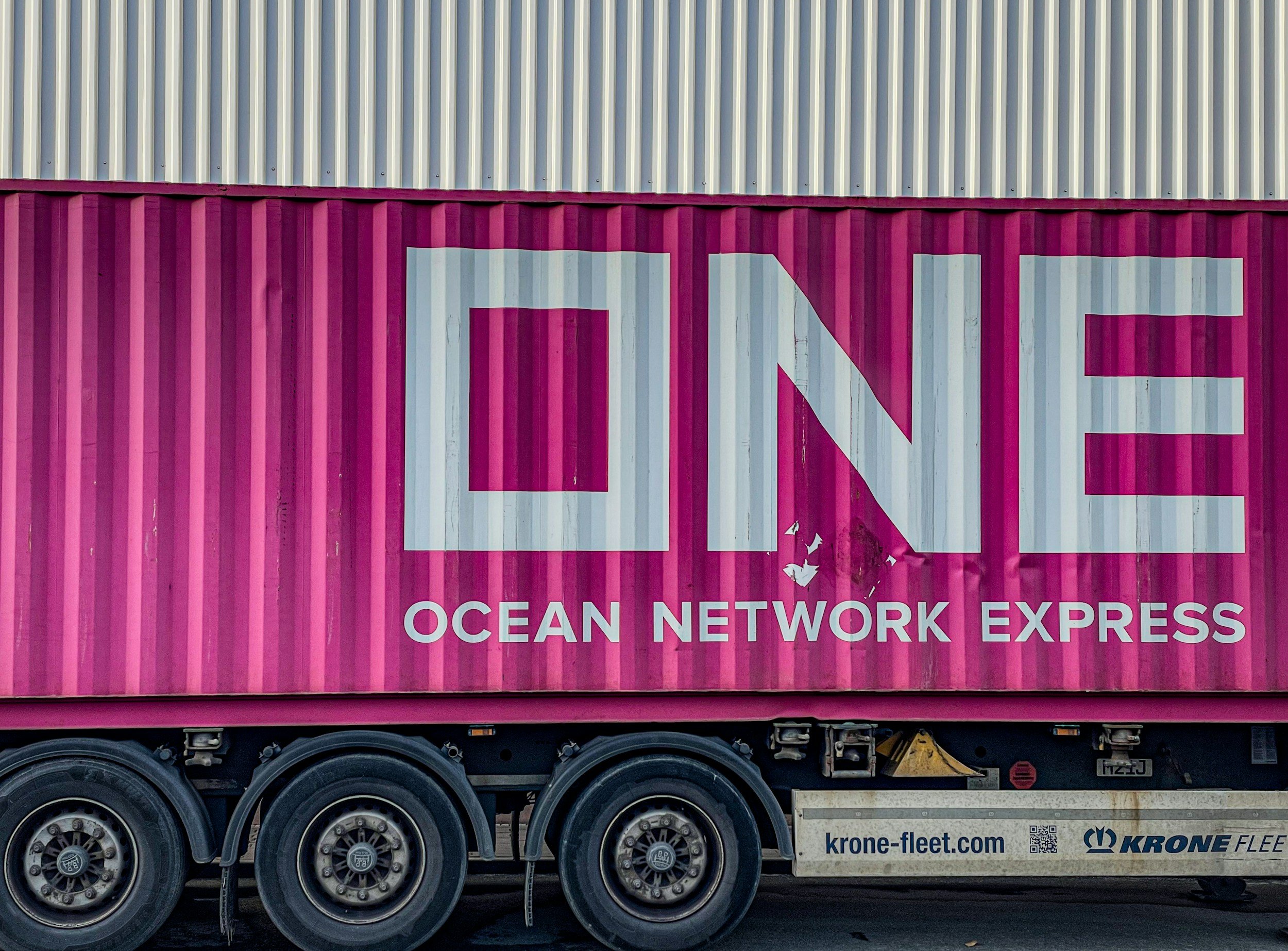 A pink shipping container, mounted on a black truck trailer with wheels delivering to Garden City.