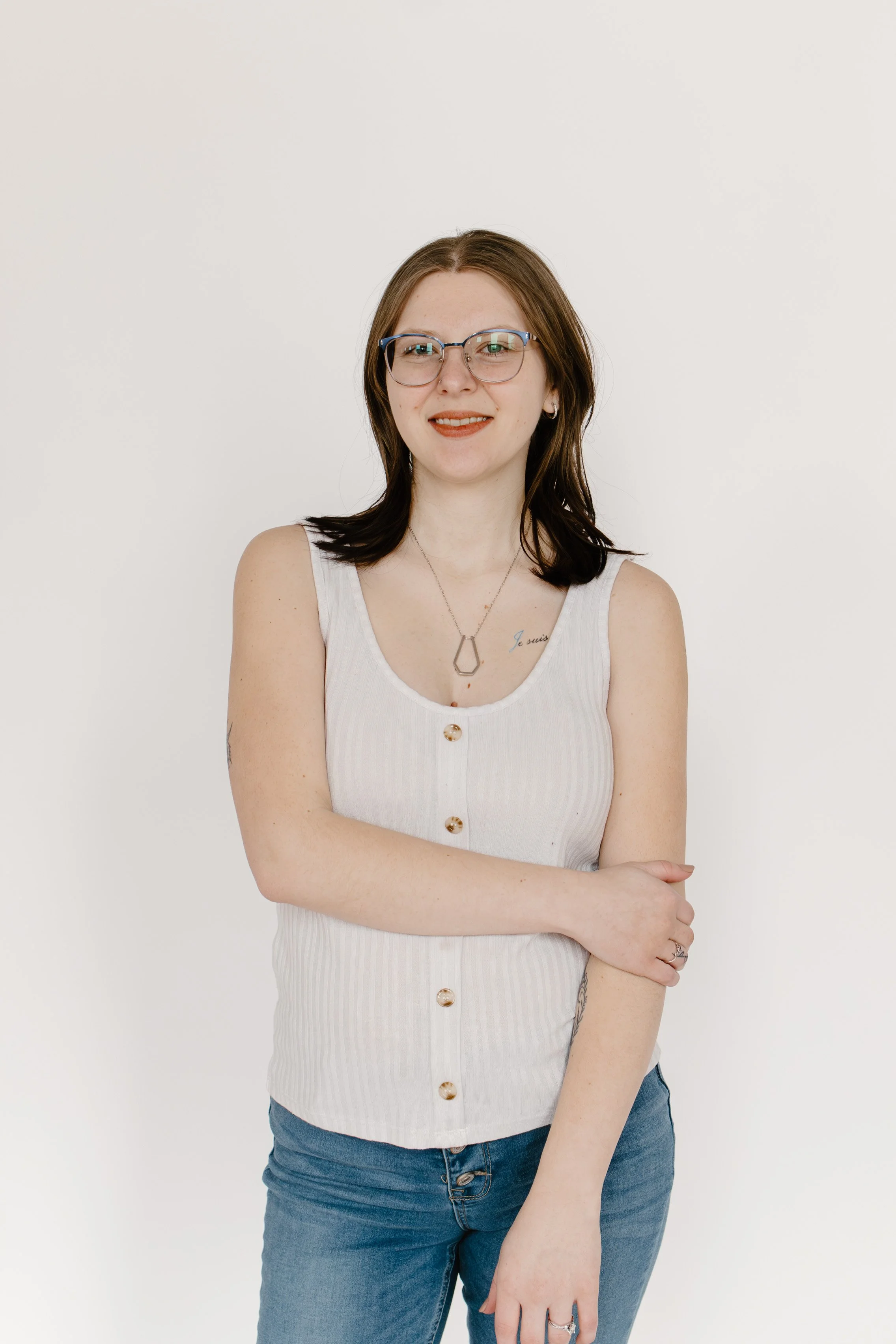 Portrait of a young woman with shoulder-length brown hair, glasses, and a tattoo on her left collarbone, standing against a plain white background. She is wearing a sleeveless white top with buttons and blue jeans, and has her arms crossed.