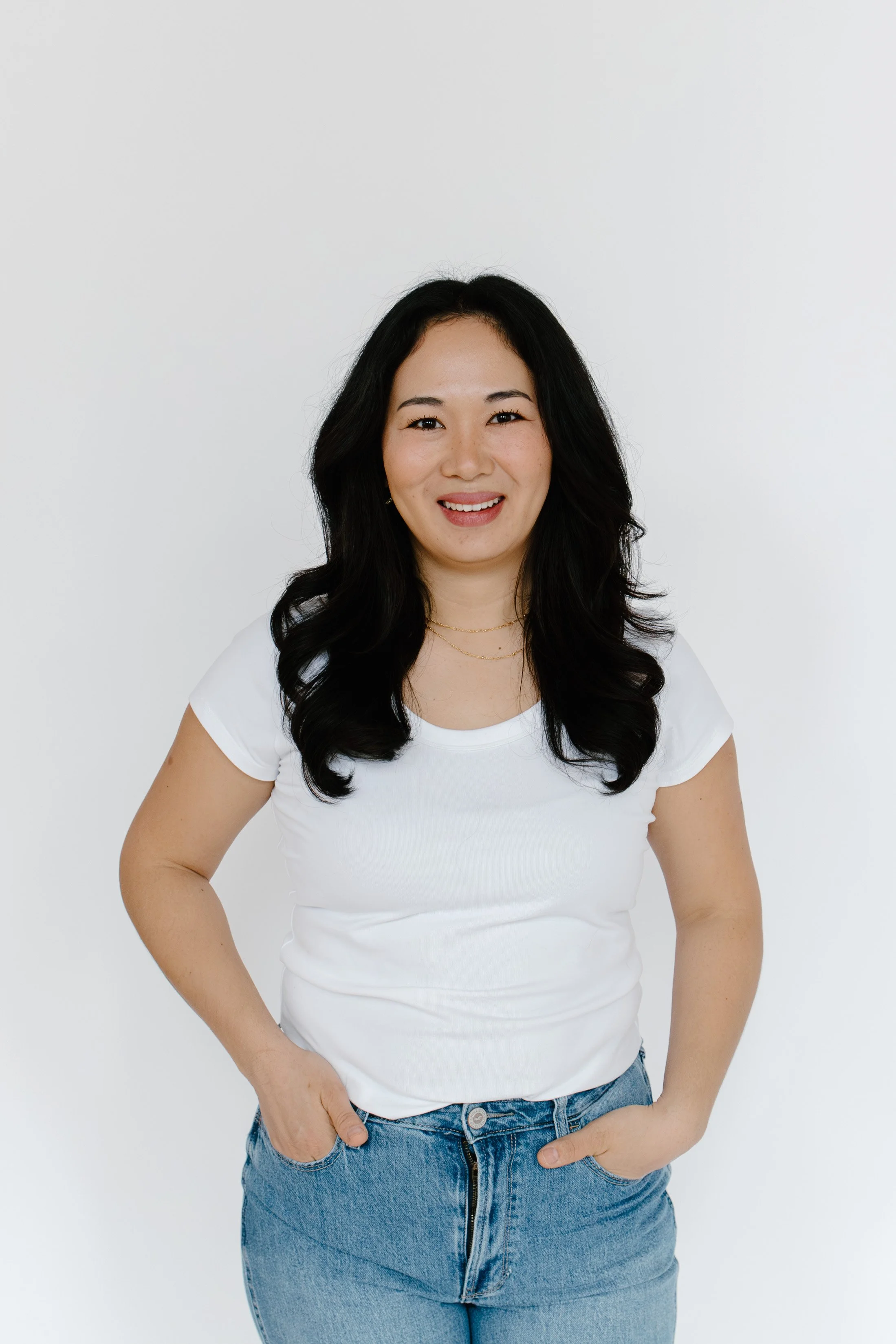 Portrait of a woman with long dark hair, wearing a white t-shirt and blue jeans, smiling and standing against a plain light-colored background.