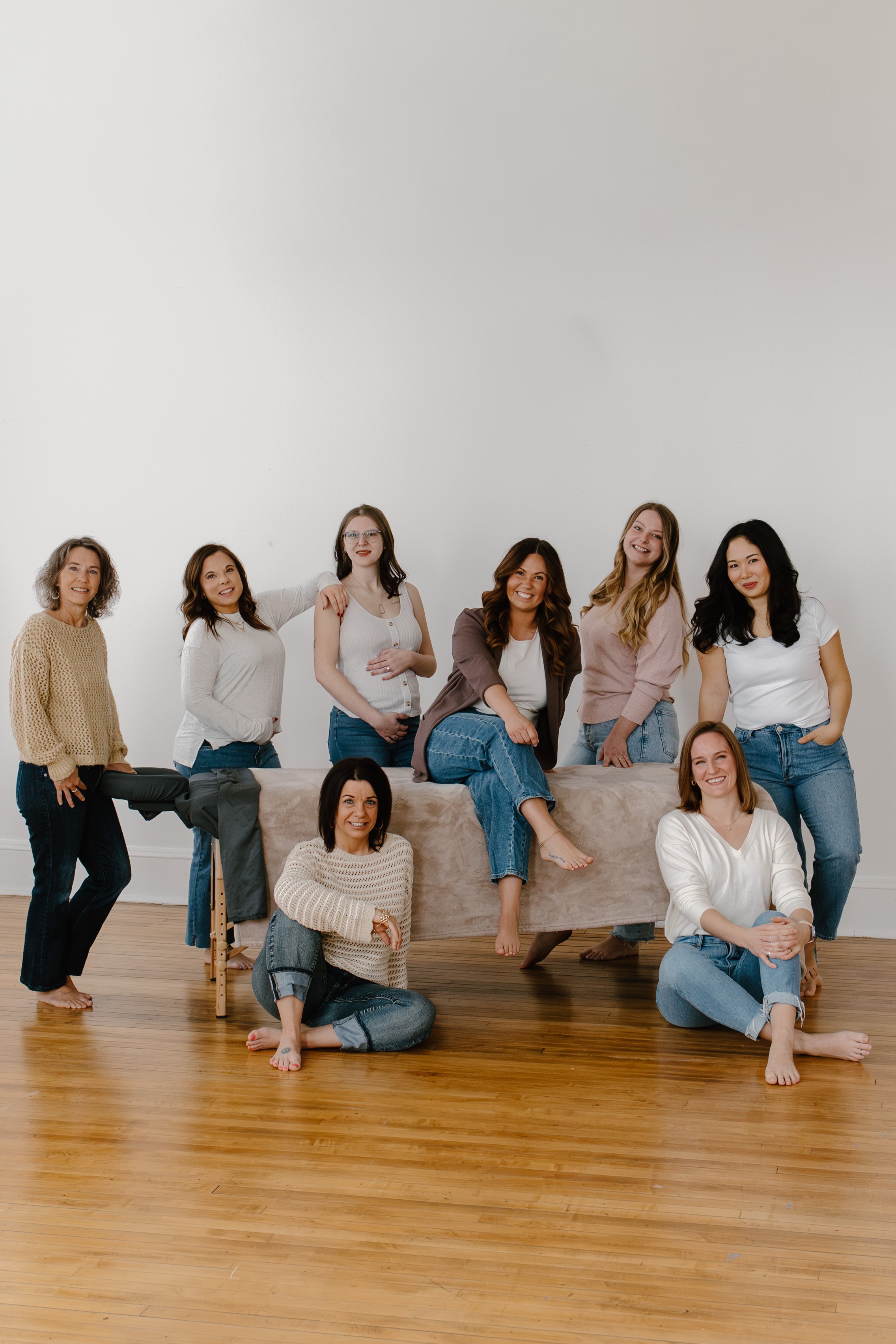 A group of nine women posing together in a bright, minimalistic room with a white wall and wooden floor. Some women are sitting on the floor, others are standing, and a few are sitting or leaning on a table covered with a beige cloth. They are smiling and appear relaxed and happy.