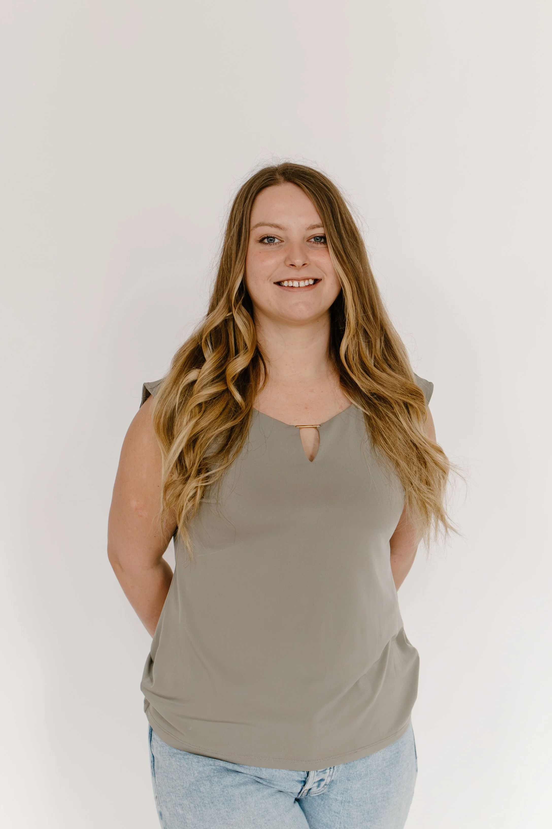 Young woman with long wavy hair wearing a taupe sleeveless top and light blue jeans, standing against a white background, smiling at the camera.