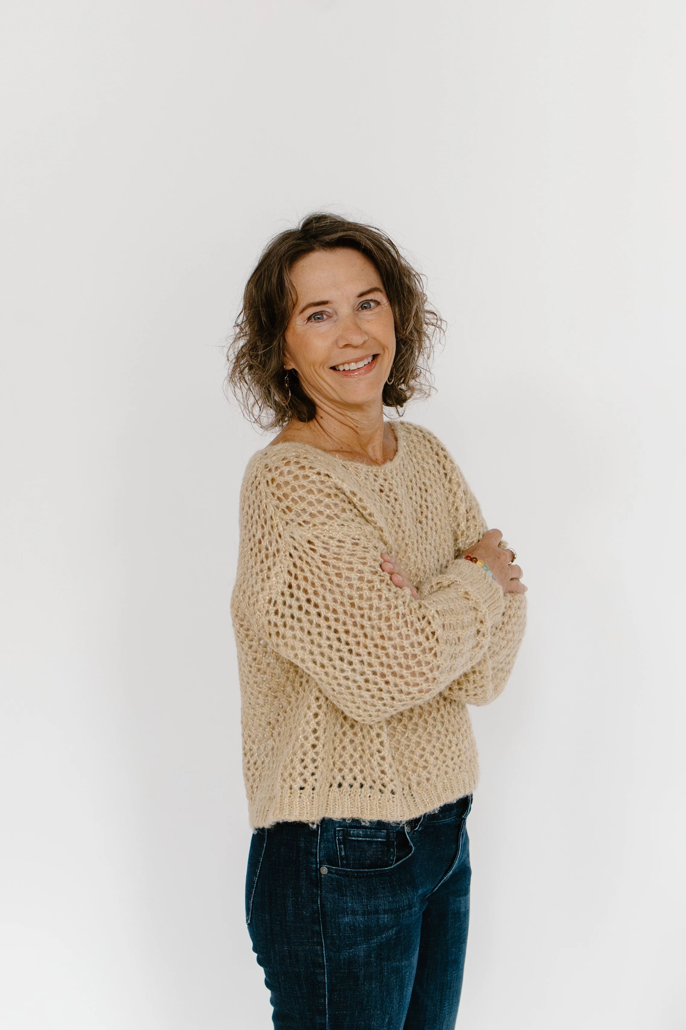 A woman with shoulder-length brown hair and a tan, open-knit sweater standing against a plain white background, smiling at the camera with arms crossed.