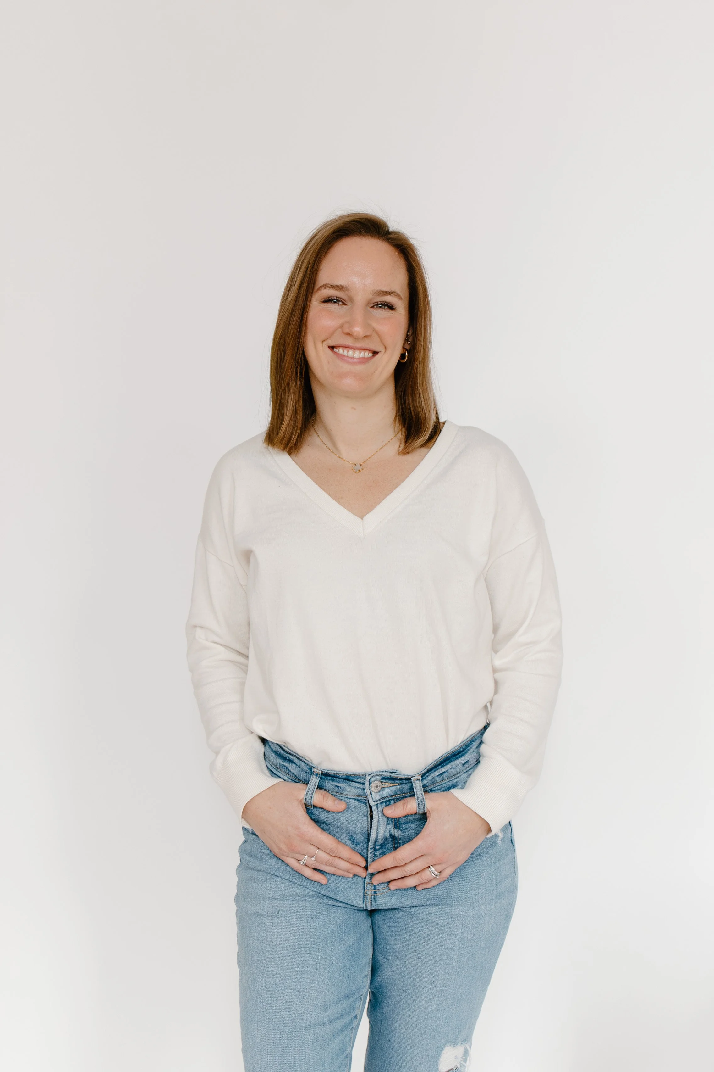A woman with shoulder-length brown hair smiling, wearing a white V-neck sweater and light blue jeans, standing against a plain white background.