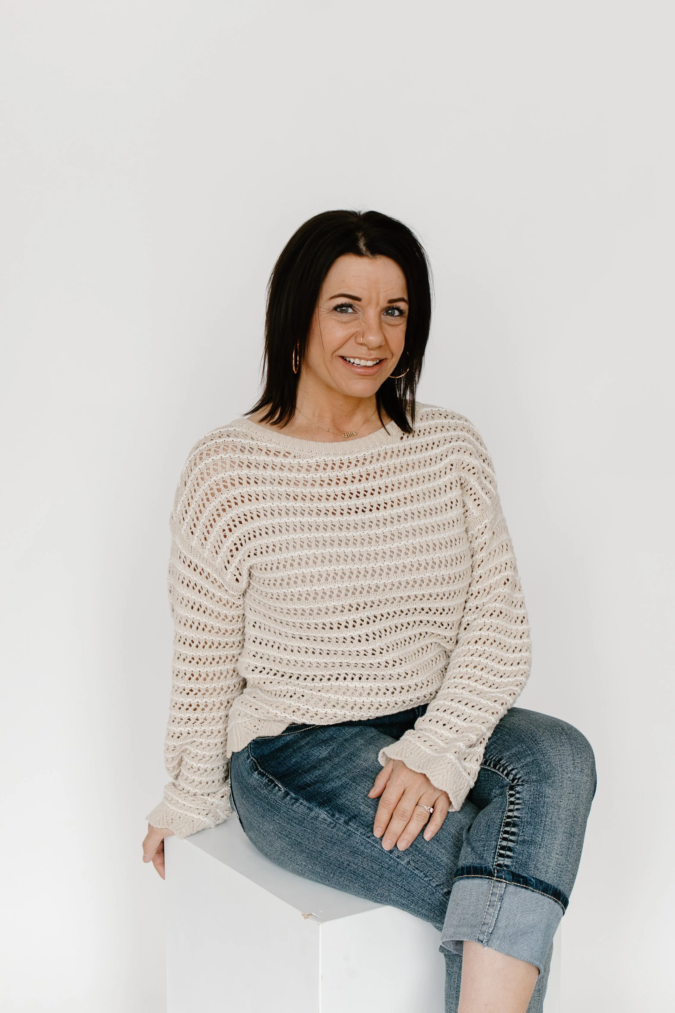 Smiling woman with black hair and hoop earrings wearing a beige open-knit sweater and blue jeans, sitting with one leg crossed on a white block against a plain white background.