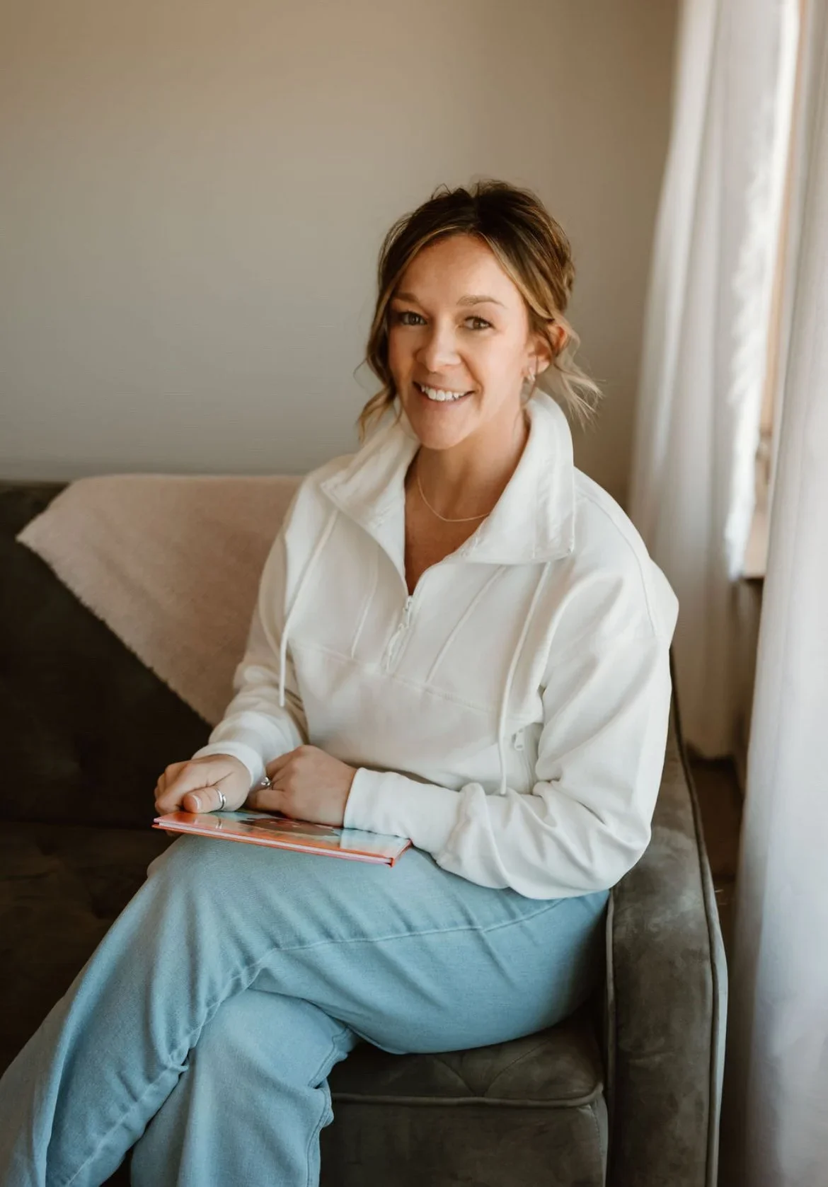 A woman with short brown hair, wearing a white zip-up jacket and light blue jeans, sitting on a couch by a window with curtains, holding a small notebook, smiling at the camera.