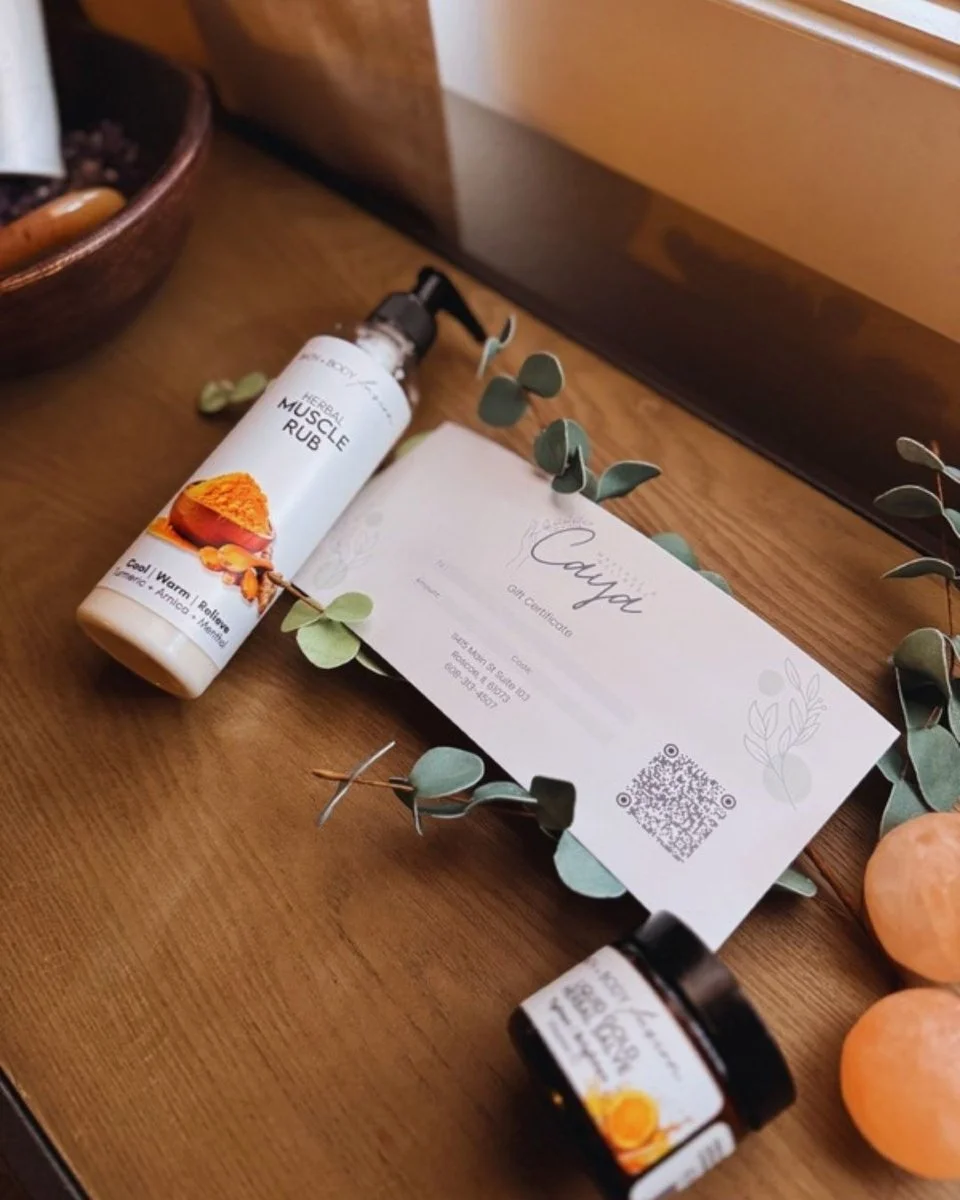 A gift certificate, a bottle of herbal muscle rub, a bottle of herbal balm, and three oranges arranged on a wooden surface with a decorative branch and a book in the background.