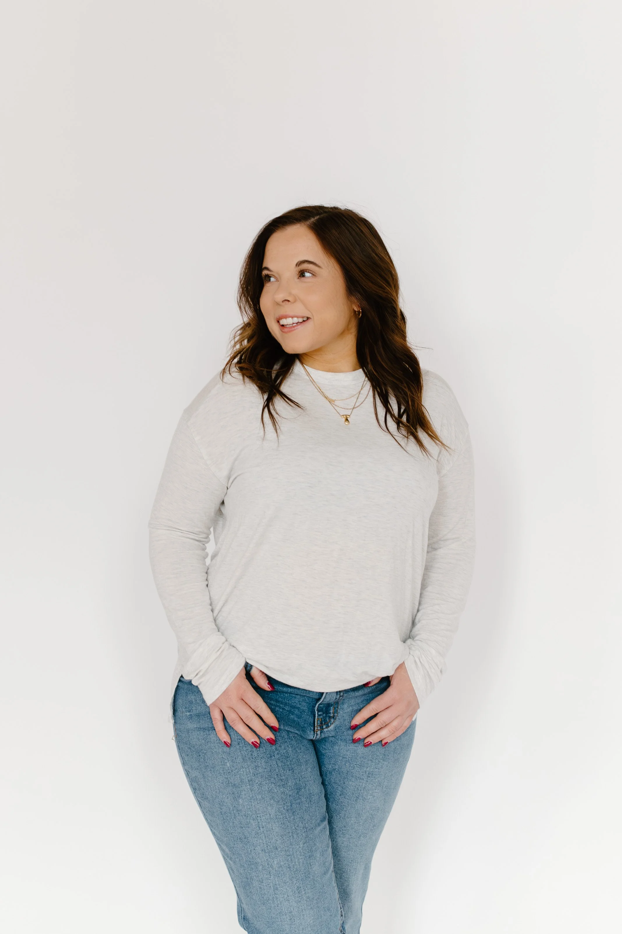 A woman with shoulder-length brown hair, wearing a light gray long-sleeve shirt, blue jeans, and red nail polish, stands against a plain white background, smiling and looking to her right.