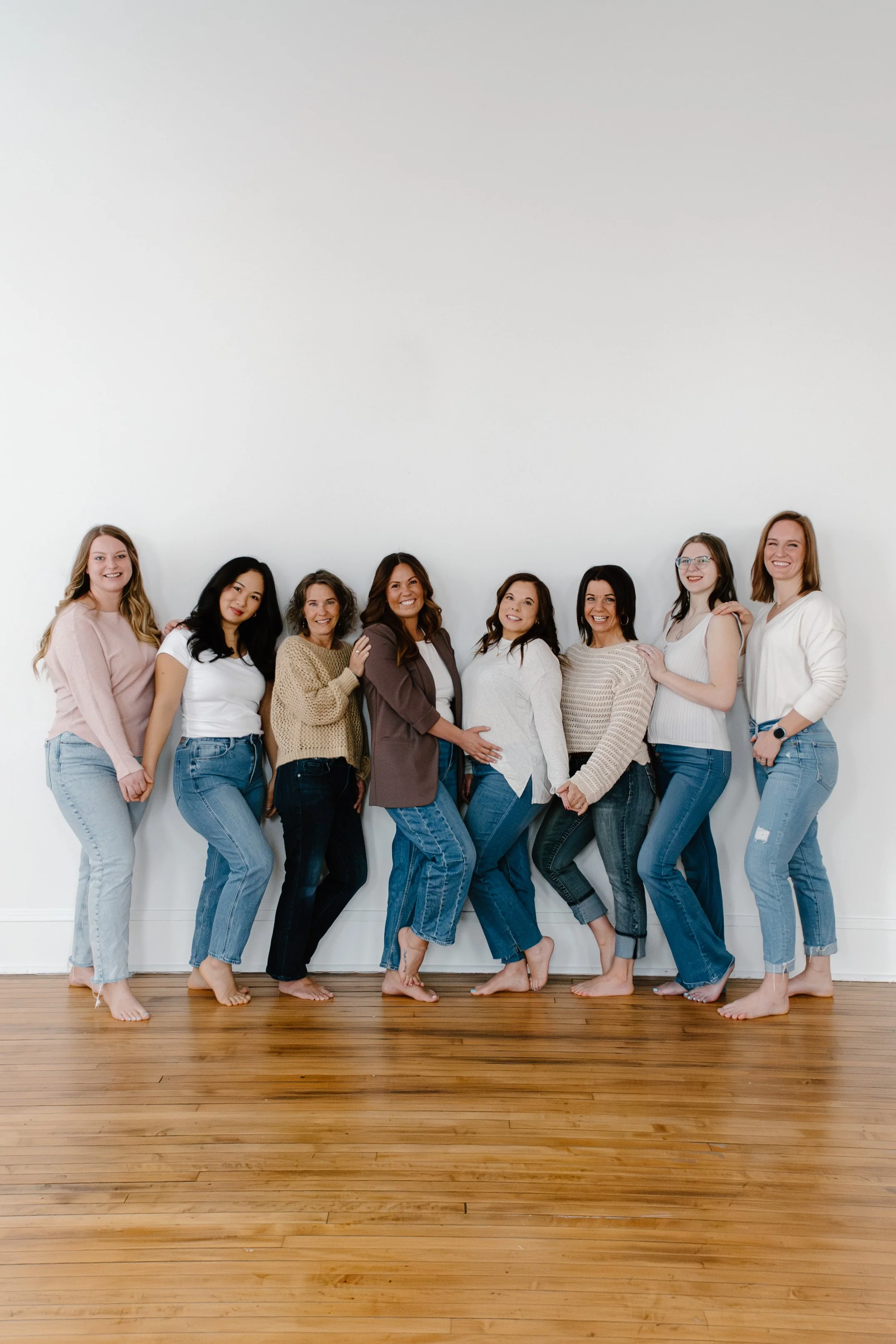 A group of nine women standing close together, smiling, and posing with their arms around each other in front of a plain white wall, all dressed casually in jeans and tops.