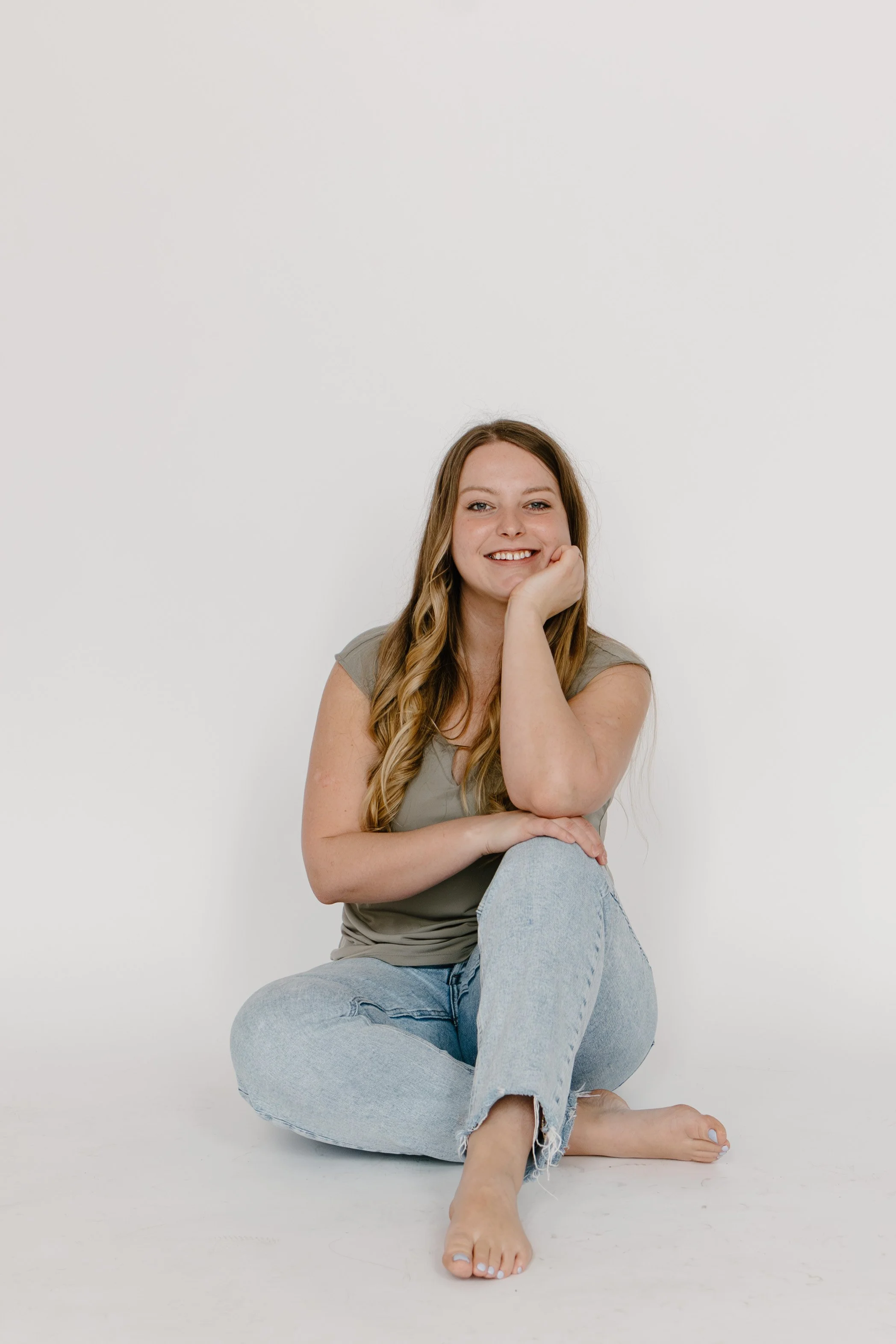 A young woman with long wavy hair, smiling, sitting on the floor with one leg crossed over the other, resting her chin on her hand, in front of a plain white background.