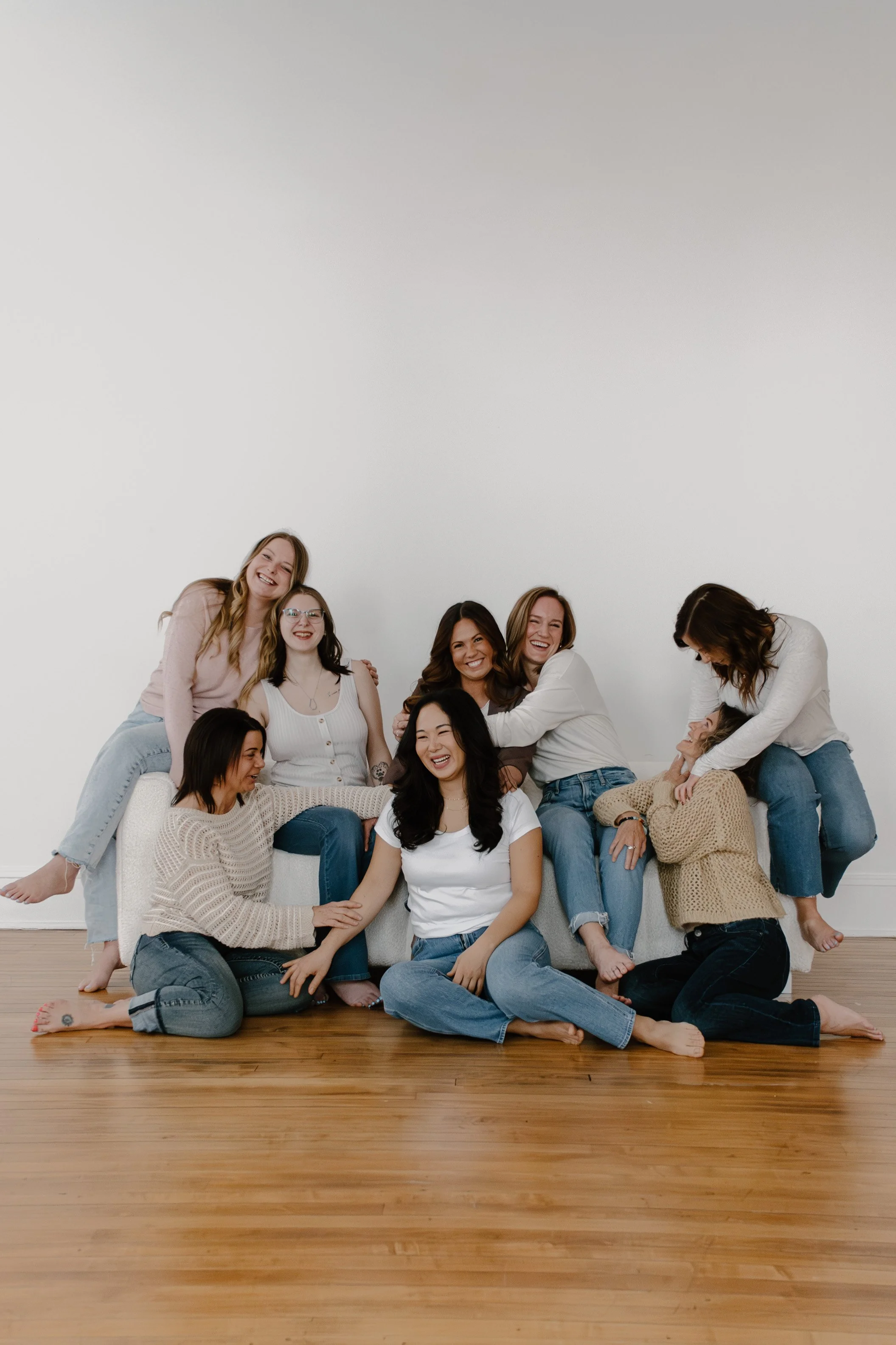Group of nine women of diverse ethnicities smiling and laughing together in a minimal white room with hardwood floors, sitting and standing on and around a beige couch.