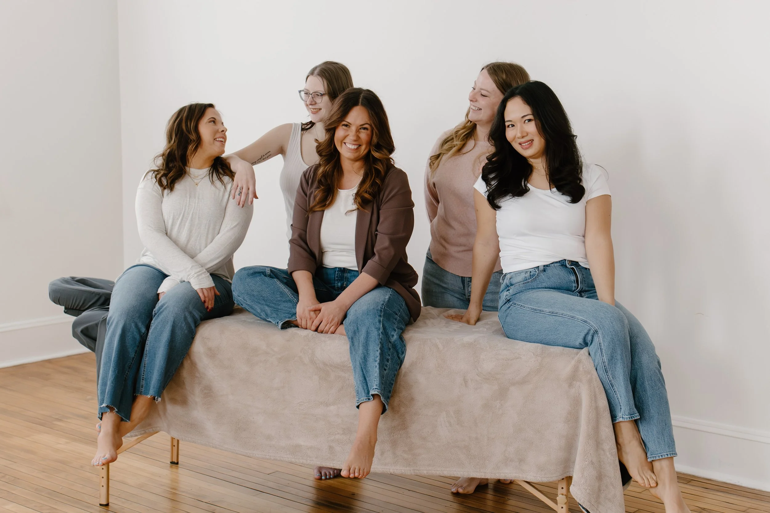 Five women sitting and standing on a massage table, smiling and laughing in a bright room.
