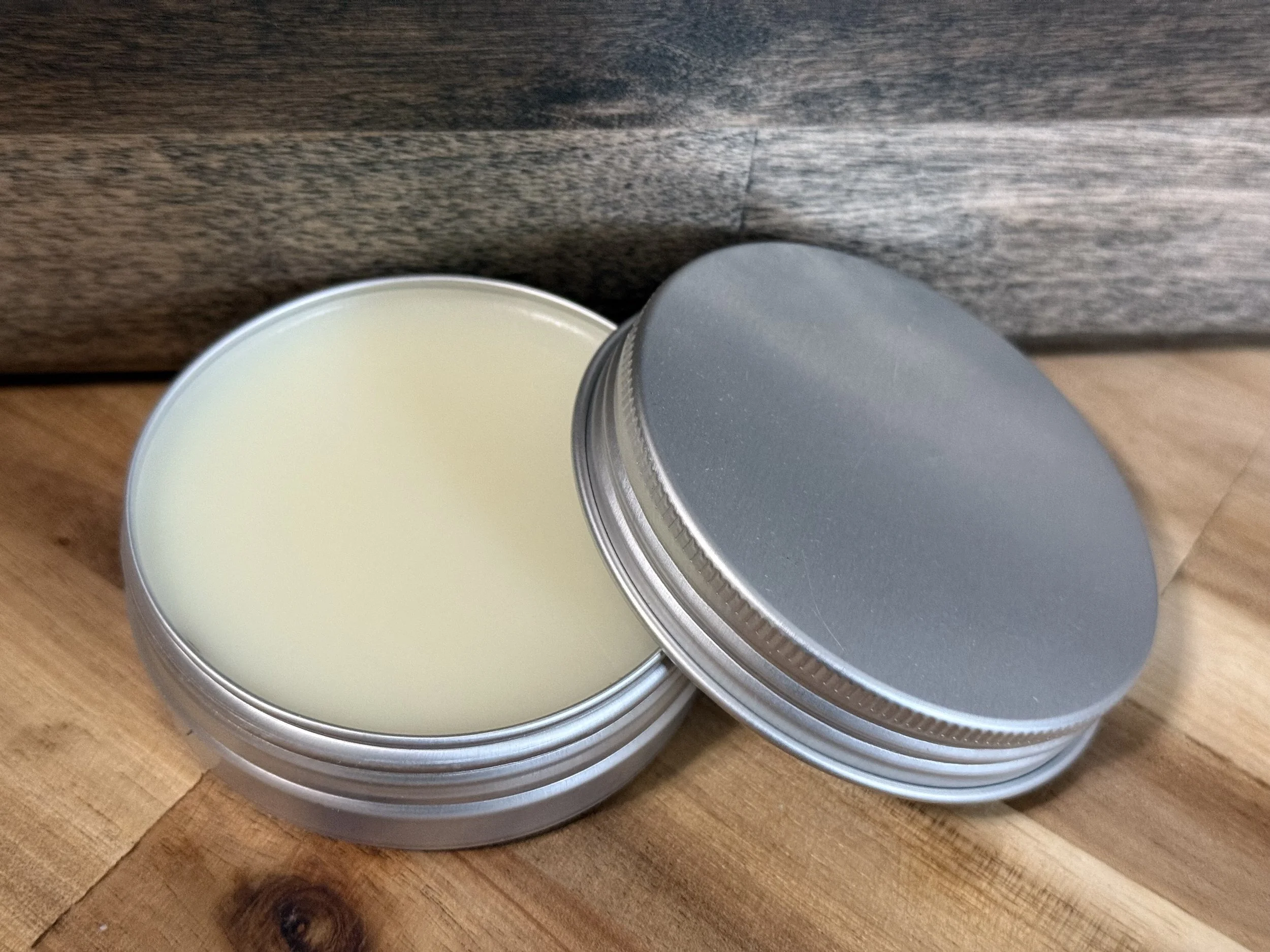 Open round metal tin container with a silver lid, filled with pale yellow wax, placed on a wooden surface against a dark wooden background.