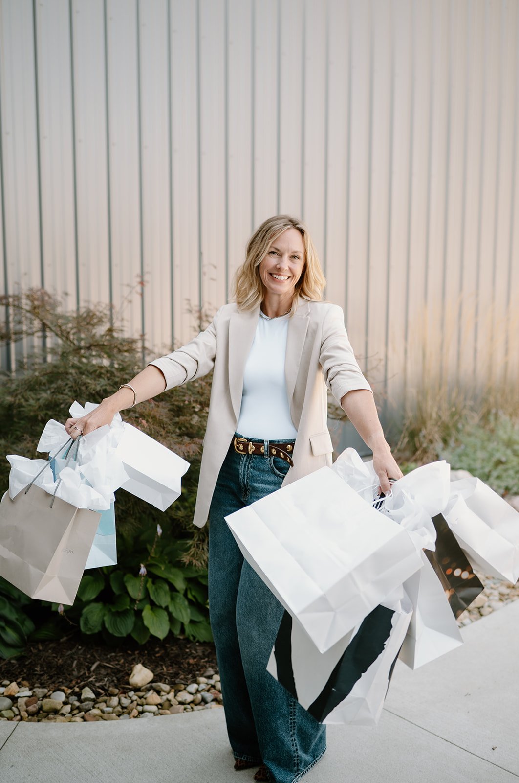 A woman with blonde hair smiling, dressed in a light blazer, white shirt, and jeans, holding shopping bags outside in front of a metallic wall.