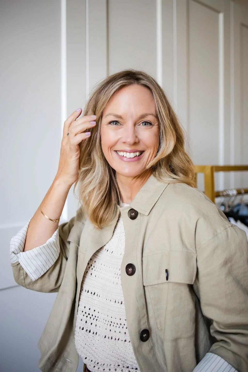 A smiling woman with shoulder-length blonde hair, wearing a beige jacket over a white crochet top, standing in front of a light-colored paneled wall.