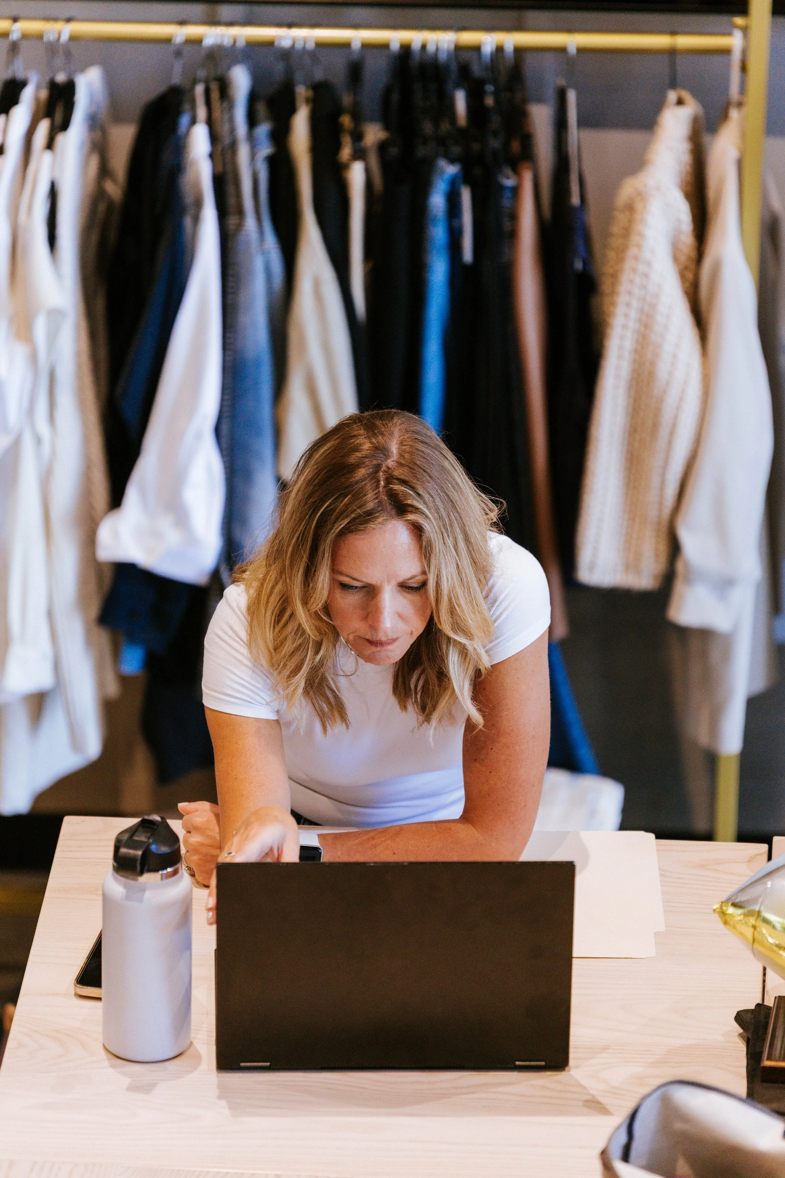 A woman with blonde hair sitting at a desk with a laptop, a water bottle, and a smartphone. Behind her are clothing racks with assorted garments.