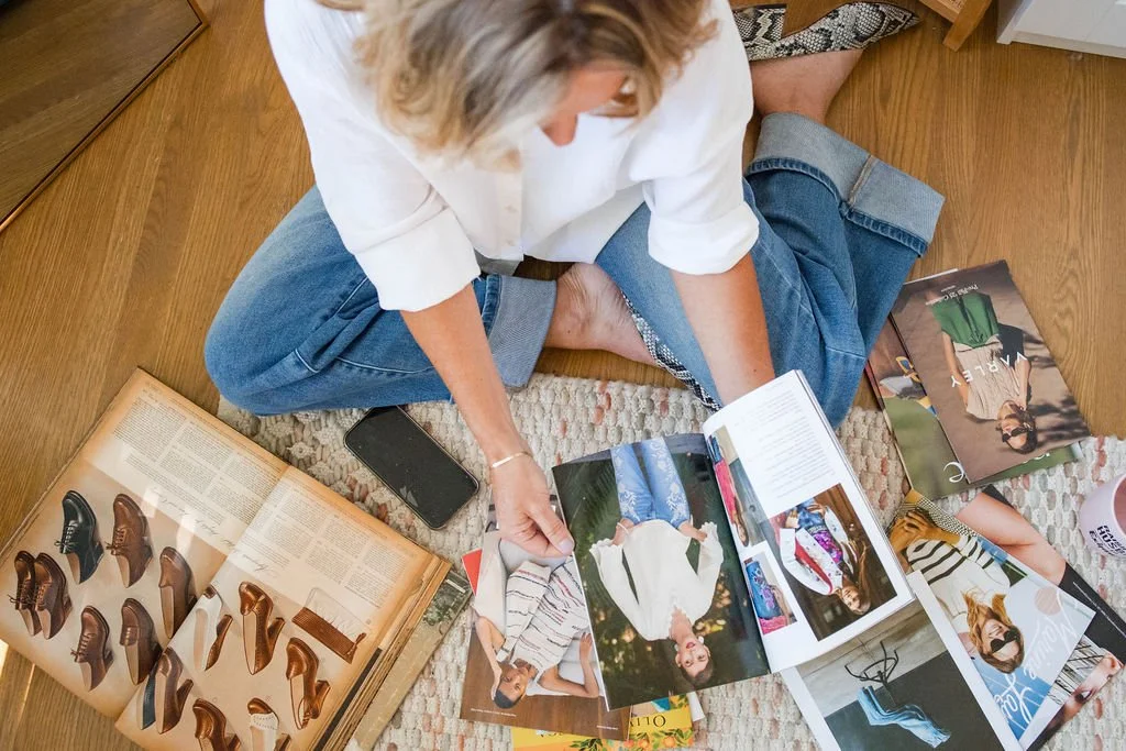 Person sitting on the floor browsing fashion magazines and catalogs, surrounded by photos of shoes and outfits, on a wooden floor with a smartphone nearby.