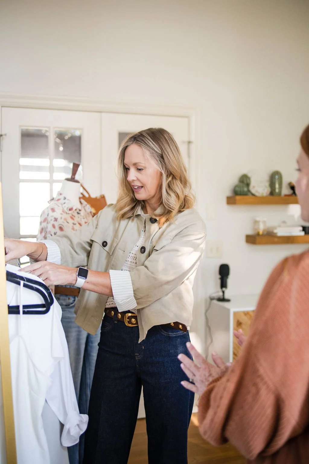 A woman with wavy blonde hair wearing a beige jacket and dark jeans is looking at clothes on a rack, showing them to a woman in a rust-colored sweater, in a well-lit room with shelves and decor in the background.