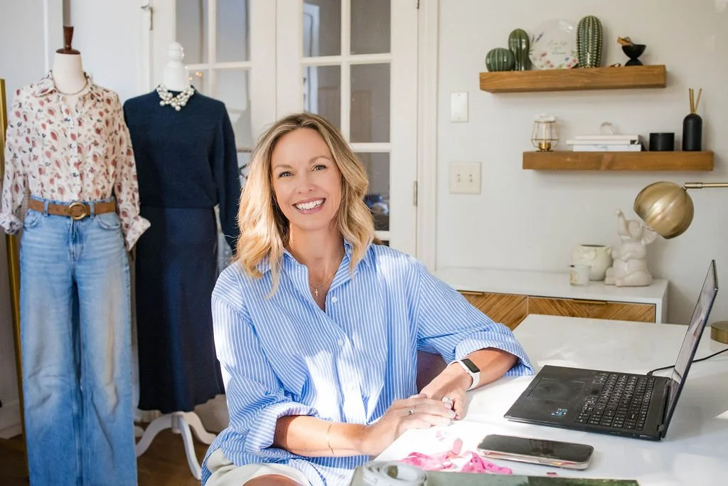 Woman sitting at a desk with a laptop and phone, smiling, in a home office with mannequins and clothing in the background, and shelves with decorative items.