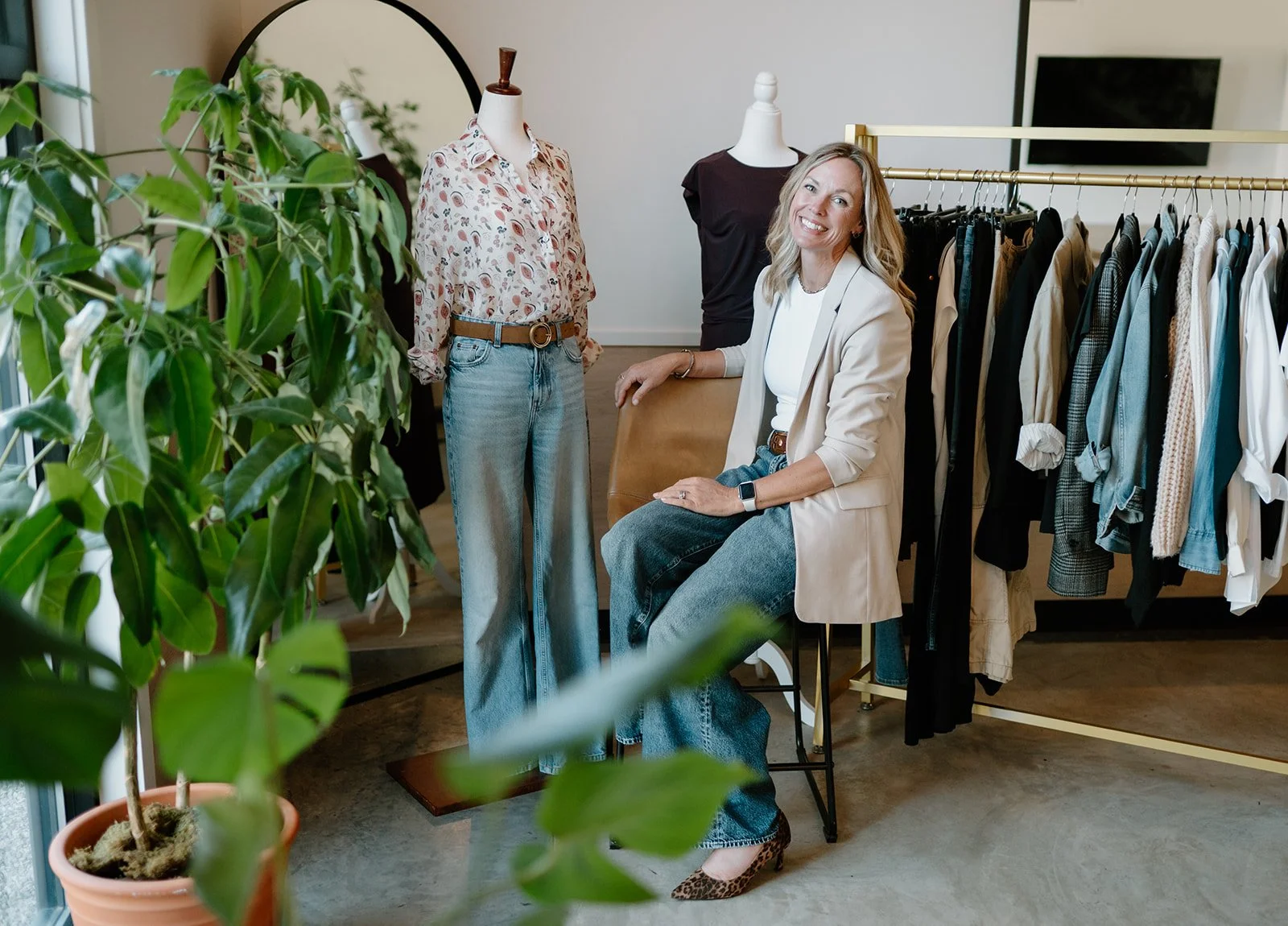 A woman with blonde hair sitting in a chair in a clothing store, smiling at the camera, surrounded by racks of clothing and mannequins, with a large green potted plant in the foreground.