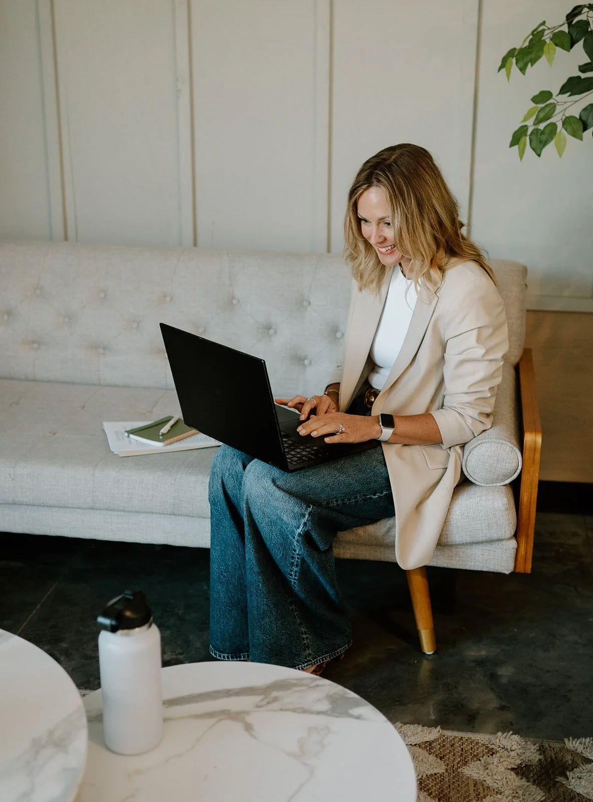 A woman sitting on a beige couch working on a black laptop, with notebooks and a pen beside her, in a room with light-colored walls and a potted plant.