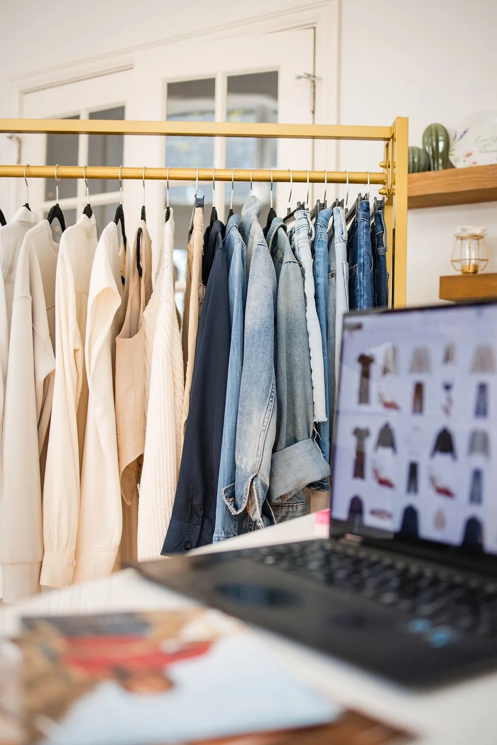 Clothes hanging on a rack with various garments including white tops, beige and cream garments, and blue jeans, with a blurred laptop and magazine in the foreground.