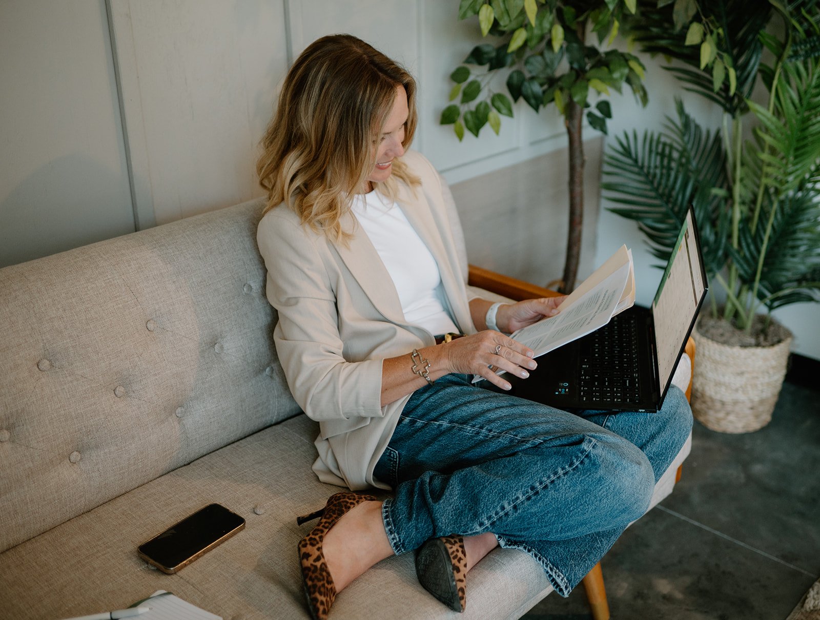 A woman with wavy blonde hair in a beige blazer and jeans, sitting on a beige sofa, reading a document, with a laptop on her lap. A smartphone is on the sofa next to her. She is wearing leopard-print high heels and a watch. Behind her are green potted plants and a white wall.