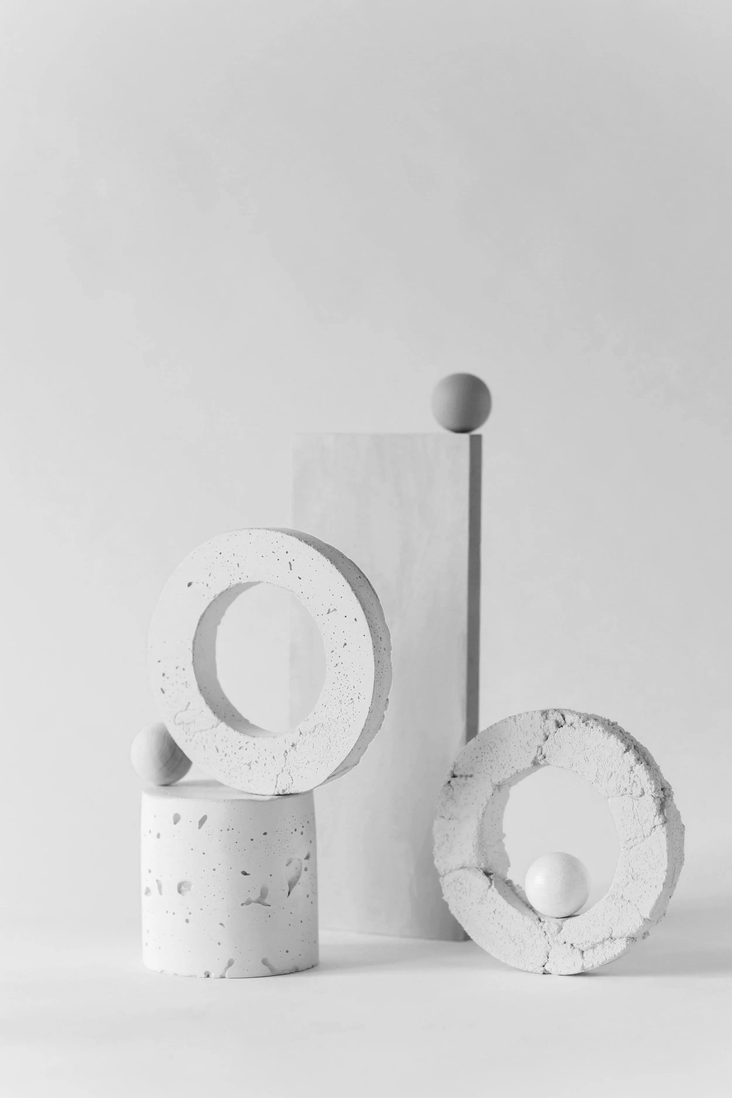 Minimalist black and white abstract arrangement with cylindrical and torus-shaped objects, including spheres and rings, on a plain background.
