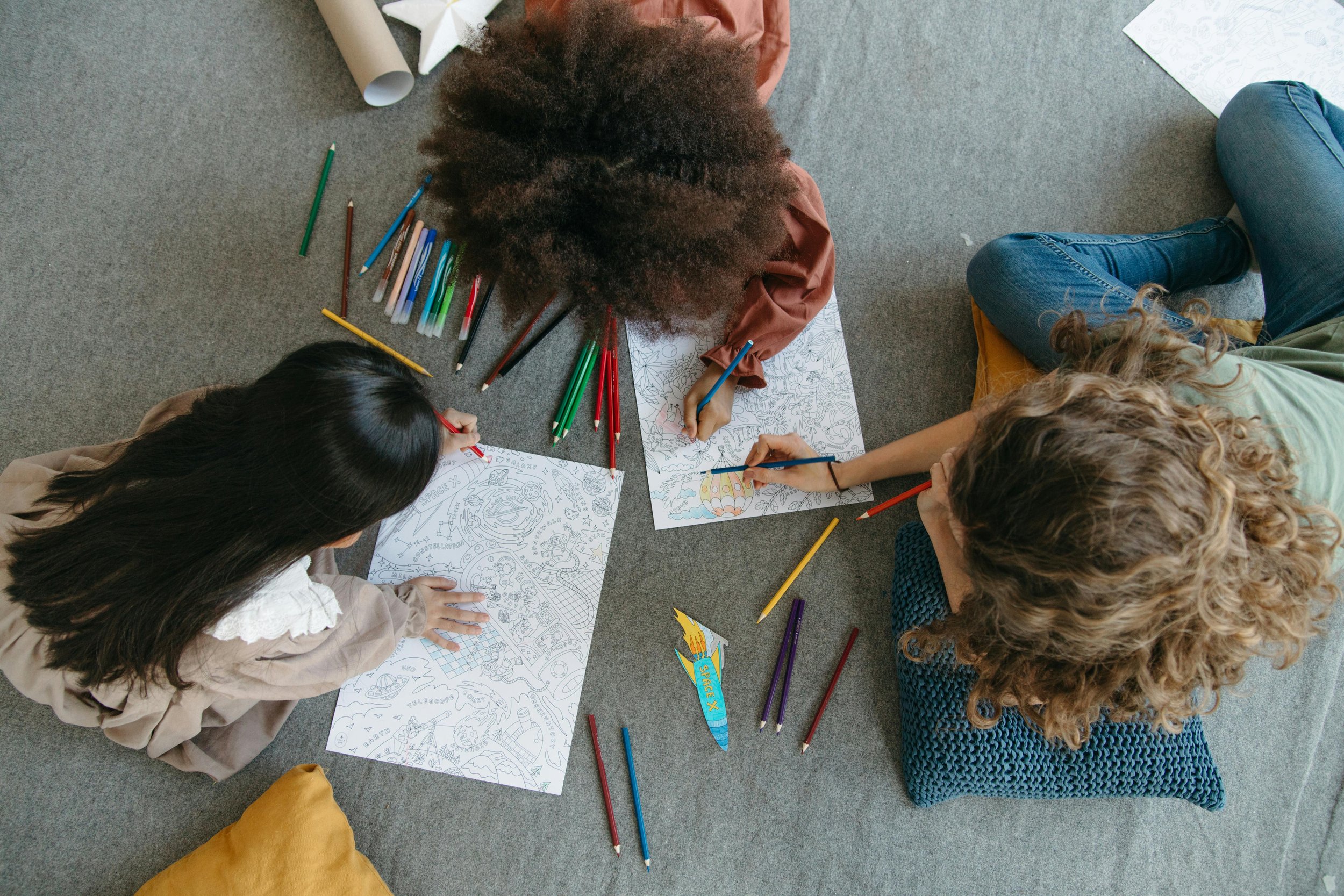 Three people lying on their stomachs on a gray carpet, coloring on large sheets of paper with various colored markers and pencils, surrounded by additional markers and pencils.